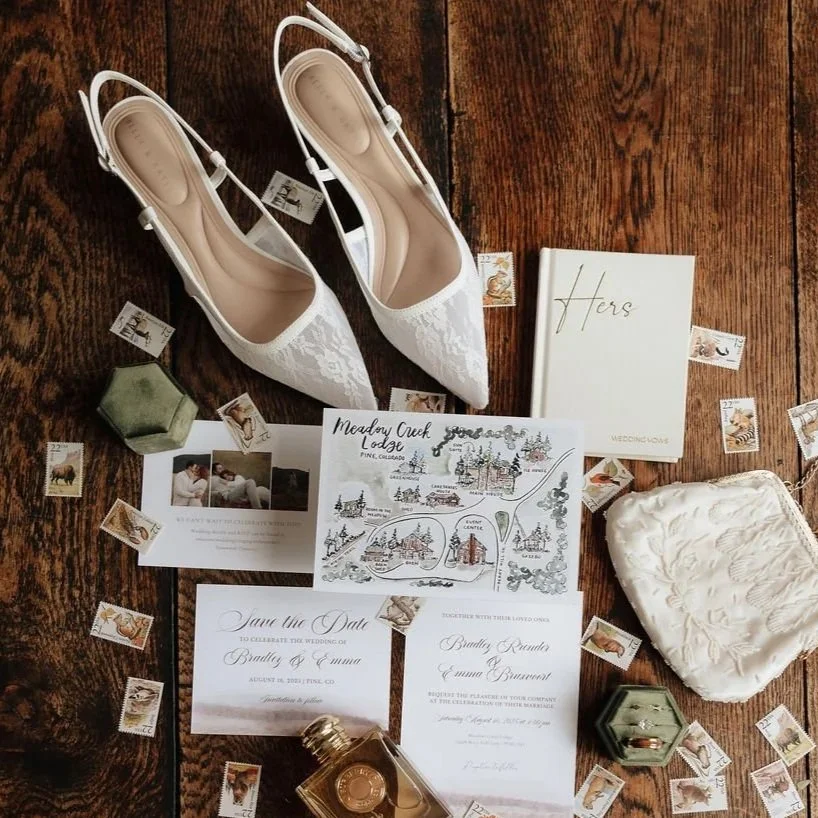 White lace high-heeled shoes surrounded by wedding invitations, a watercolor map of Meadow Creek Lodge, stamps, and a small jewelry box on a wooden surface.