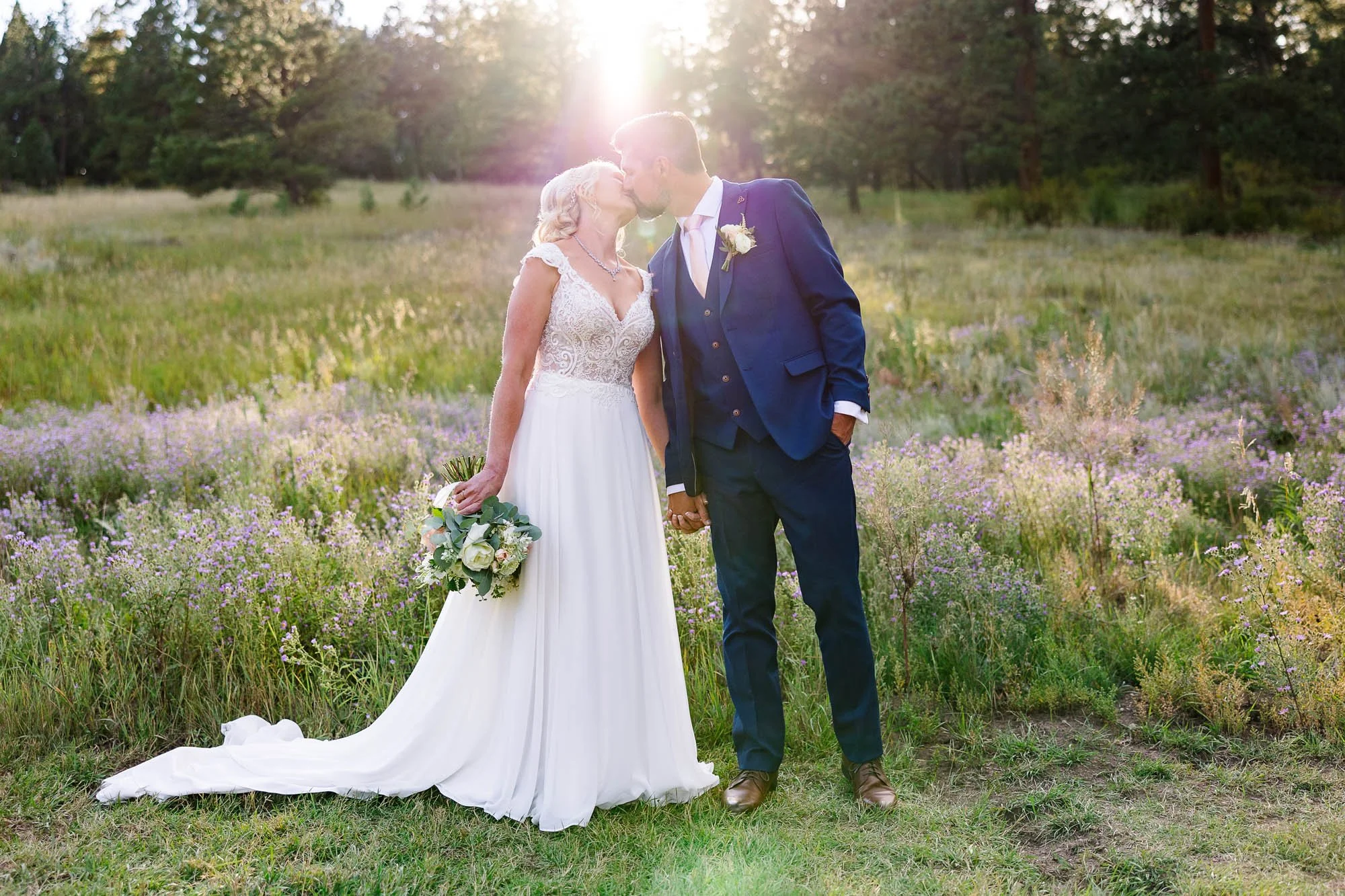 A bride and groom sharing a kiss in a field with wildflowers and trees in the background, sunlight shining behind them.