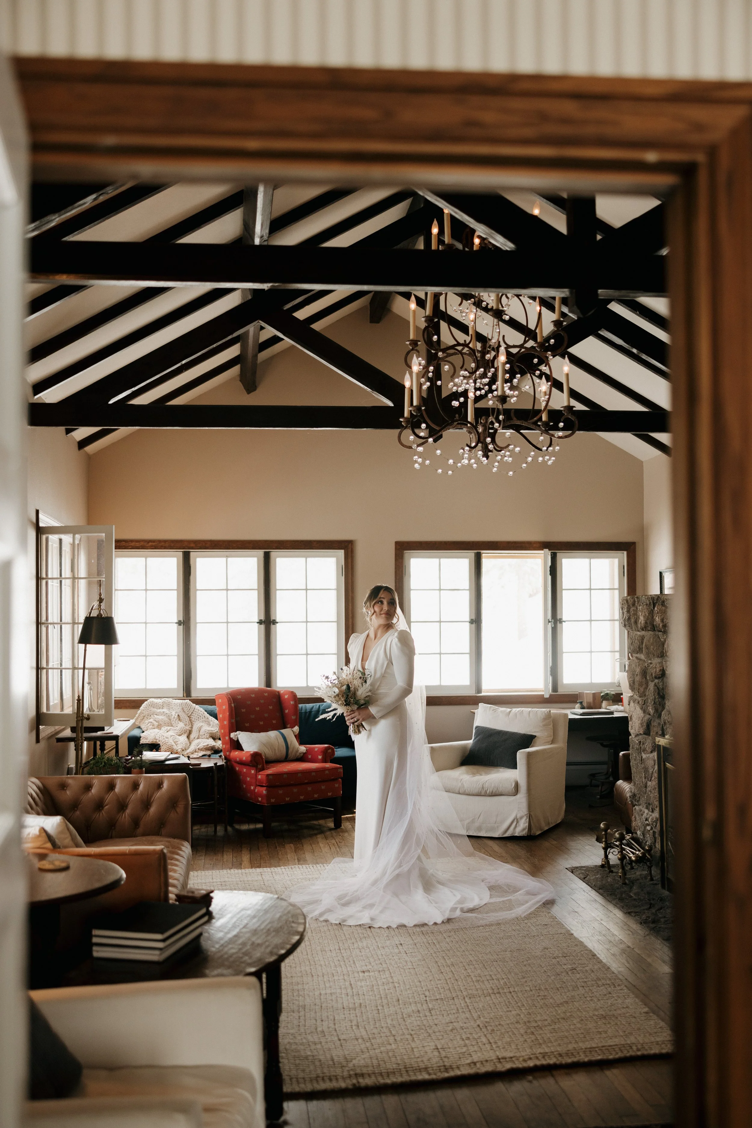 A bride in a white wedding dress holding a bouquet in a cozy, rustic living room with large windows, antique chandelier, and various chairs and couches.