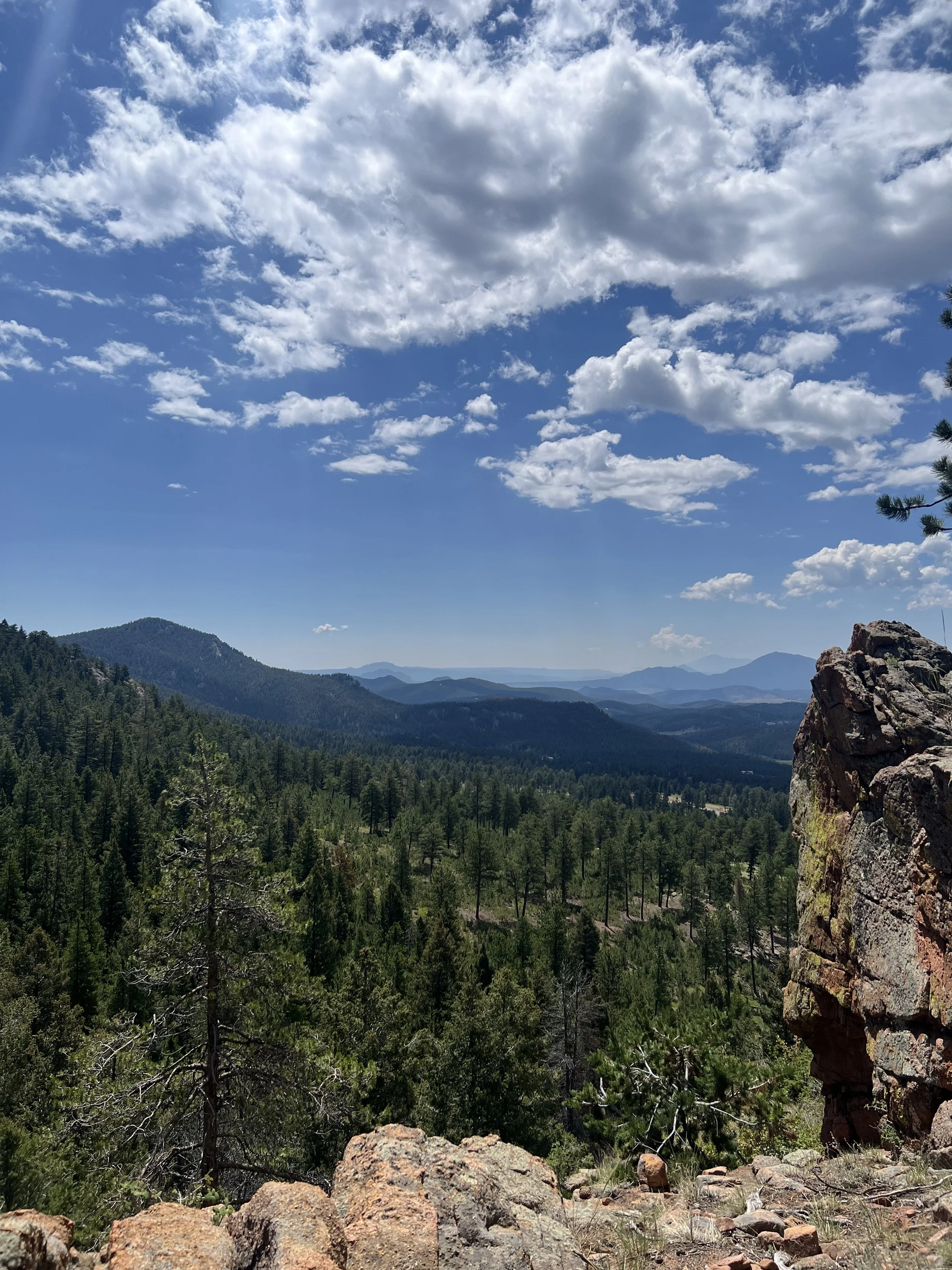 A scenic view from Staunton State Park in Pine, Colorado with forest of pine trees, a partly cloudy sky, and rocky foreground.