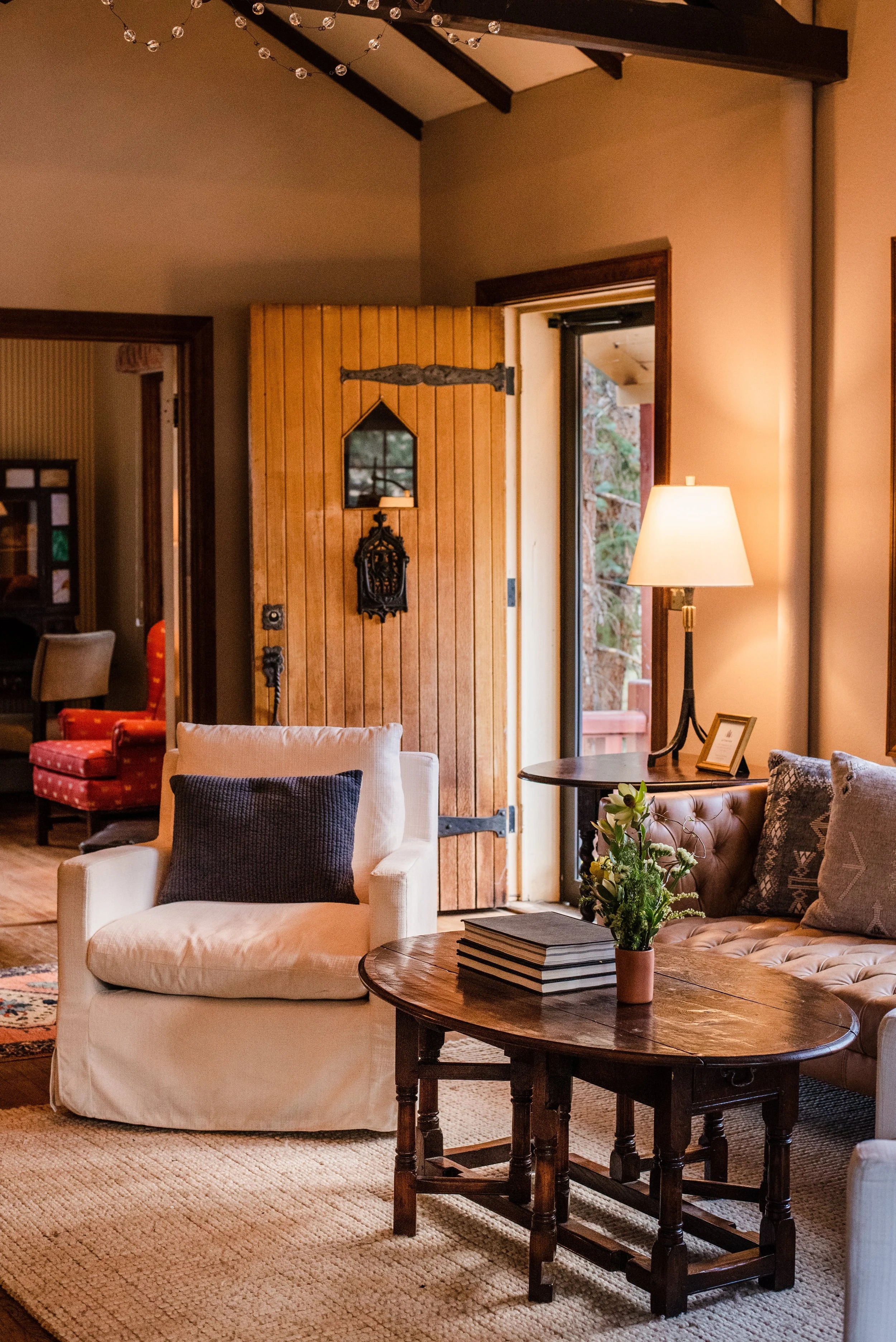 Living room with beige armchair, pillow, round wooden table with books and a plant, sofa with various pillows, a floor lamp, wooden wall with mirror, and open door.