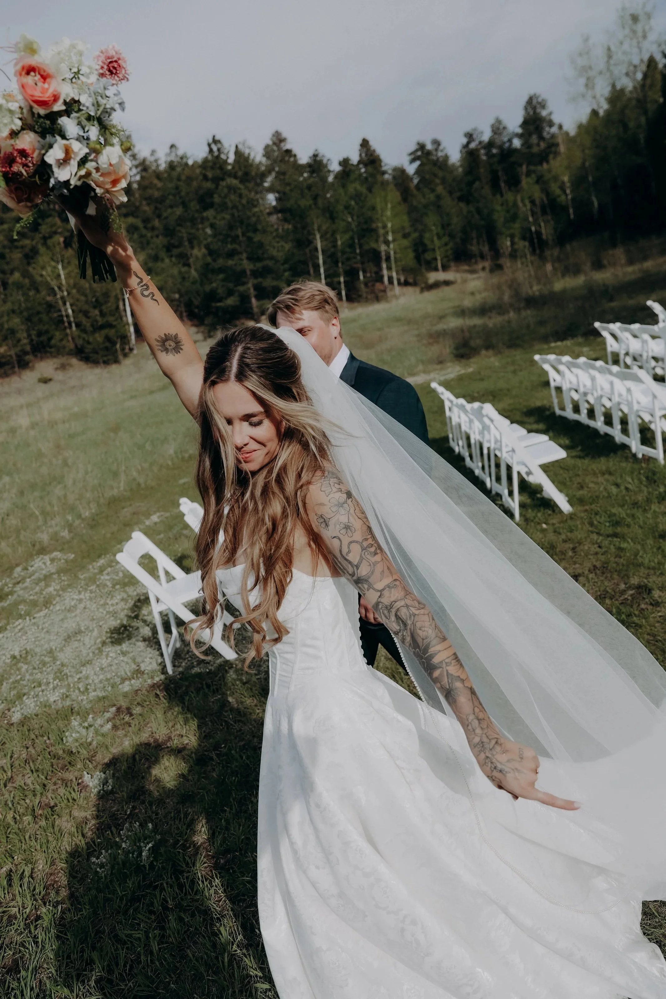 A bride holding a bouquet of flowers with one arm raised, standing outdoors in a grassy area with white chairs and trees in the background.