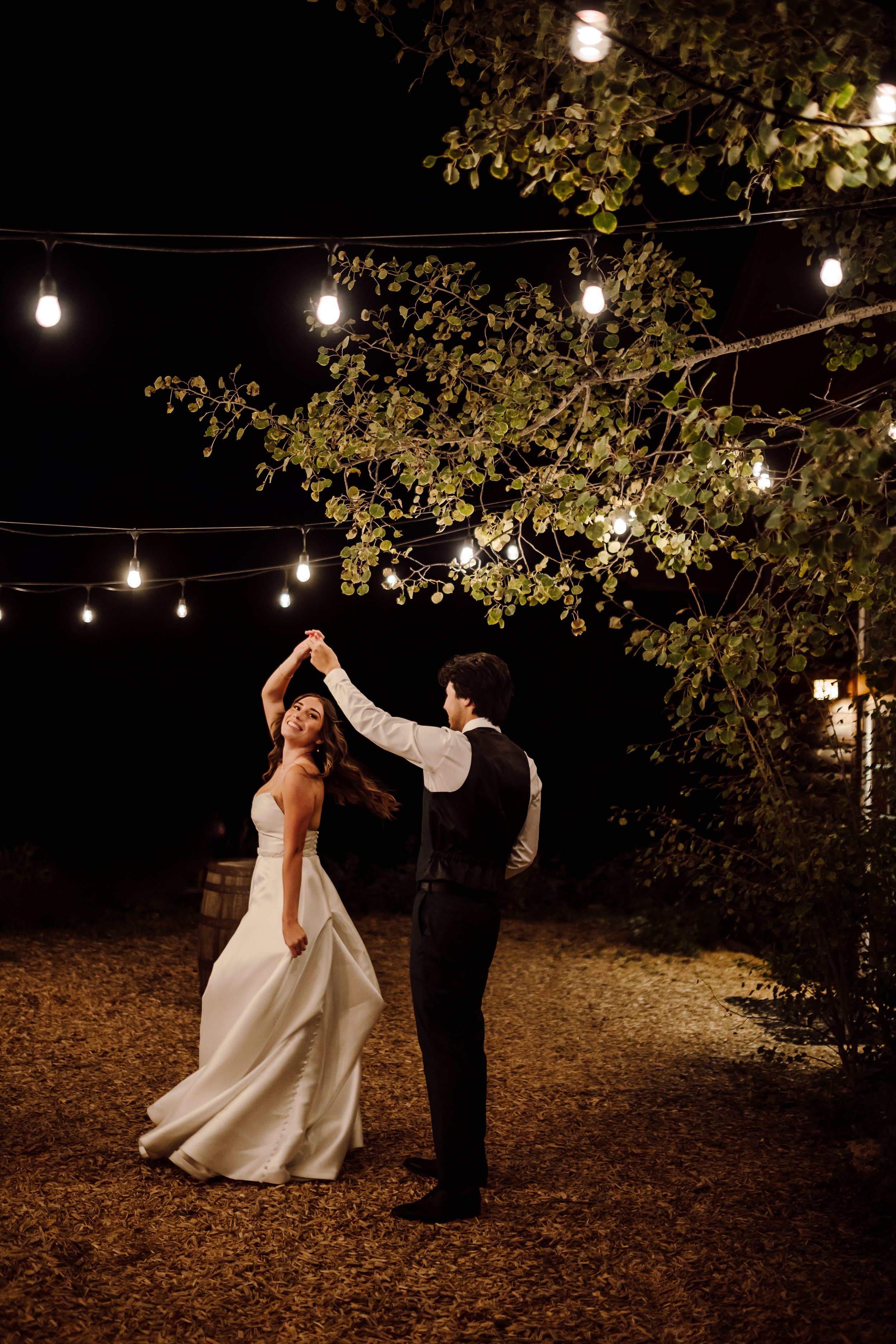 A bride and groom dancing outdoors at night under string lights, surrounded by trees with green leaves and a dark sky.