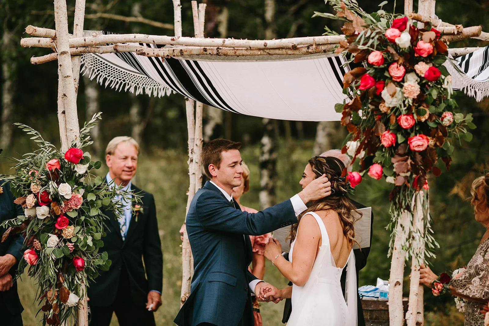 A couple getting married outdoors under a decorated canopy, with the groom tucking the bride's hair behind her ear, surrounded by friends and family.