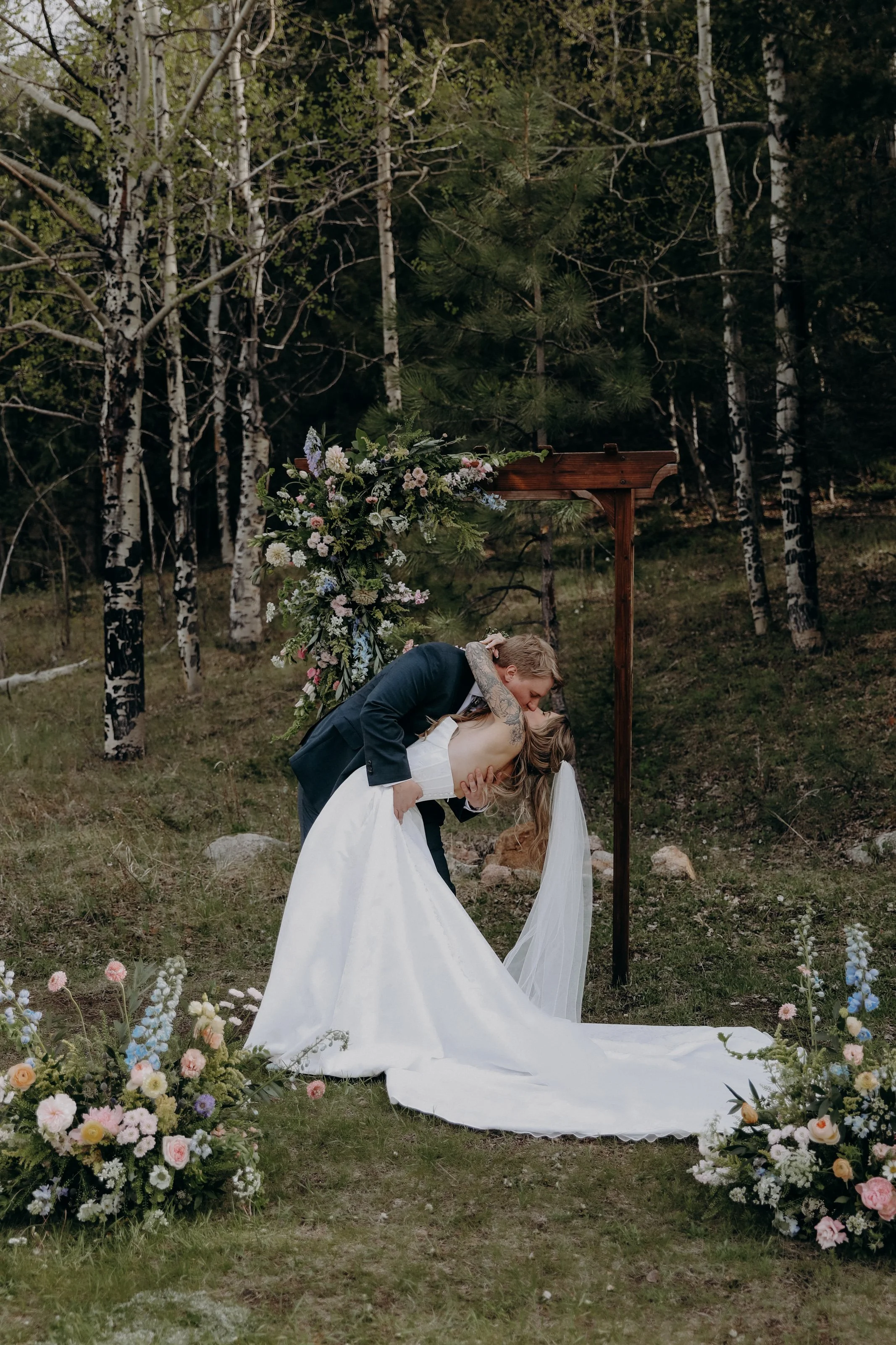A newly married couple kissing during an outdoor wedding ceremony in a wooded area decorated with flowers and a wooden arch.