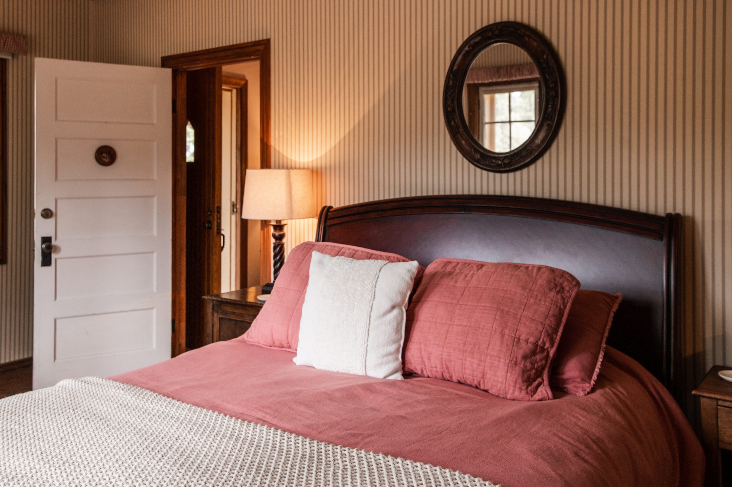 A bedroom with a wooden bed frame, pink bedding, white and pink pillows, a bedside table with a lamp, a large round mirror on striped wallpaper, an open door with a wooden frame, and a window reflected in the mirror.