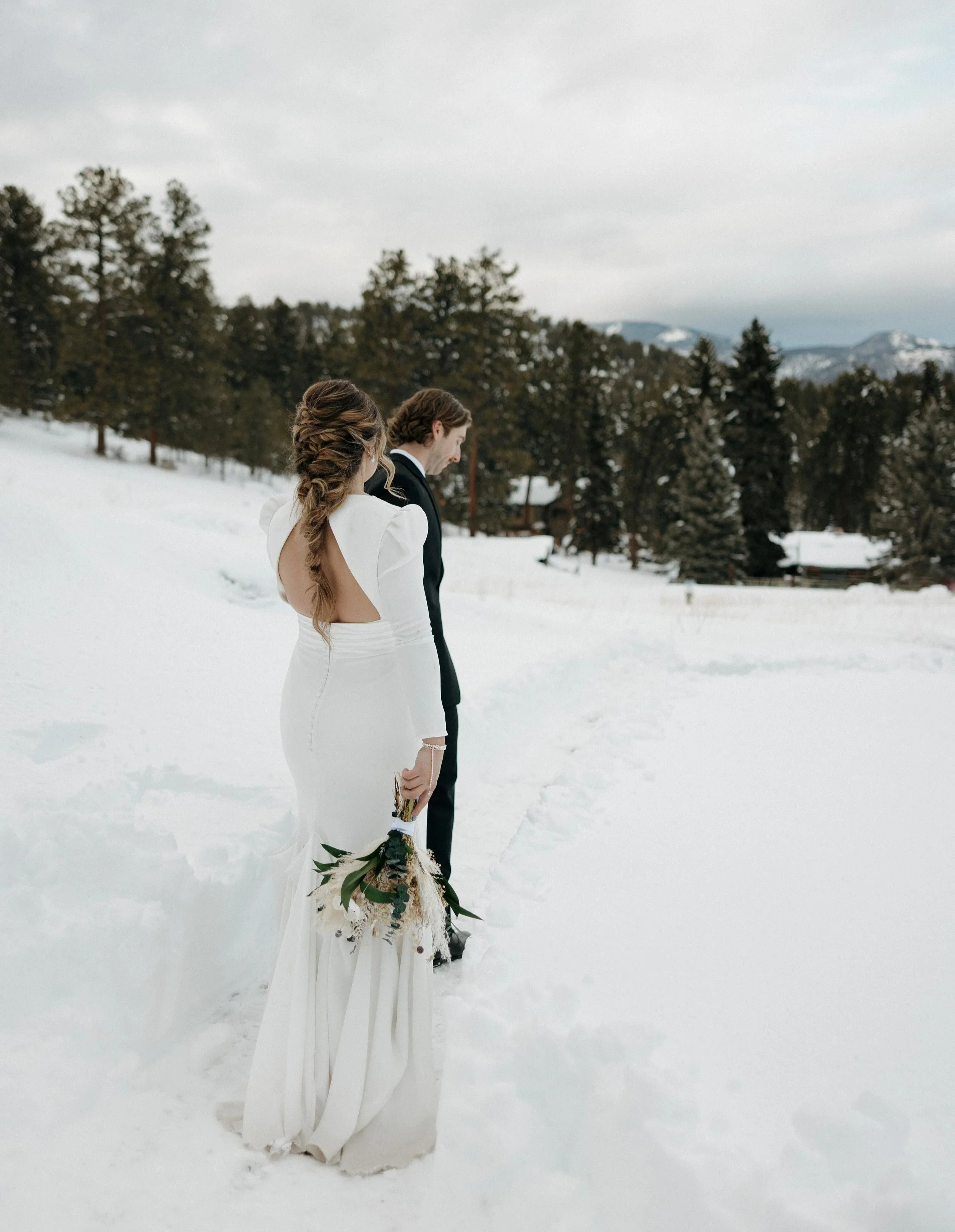 A bride and groom standing outdoors in a snowy landscape, with evergreen trees and distant mountains in the background, during winter.