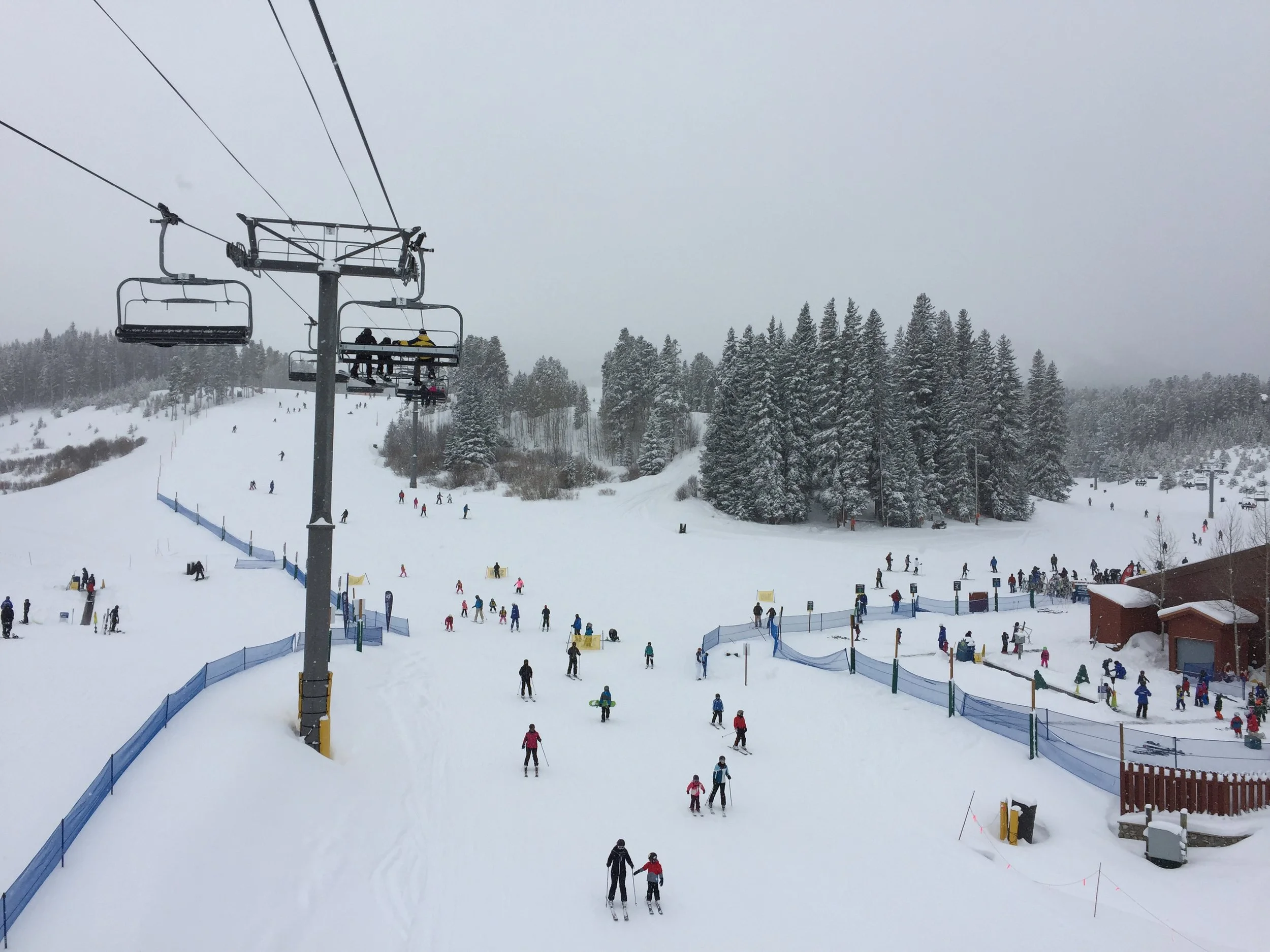 Breckenridge ski resort with snow-covered slopes, ski lifts, and multiple skiers and snowboarders. Forested hills in the background and overcast sky.