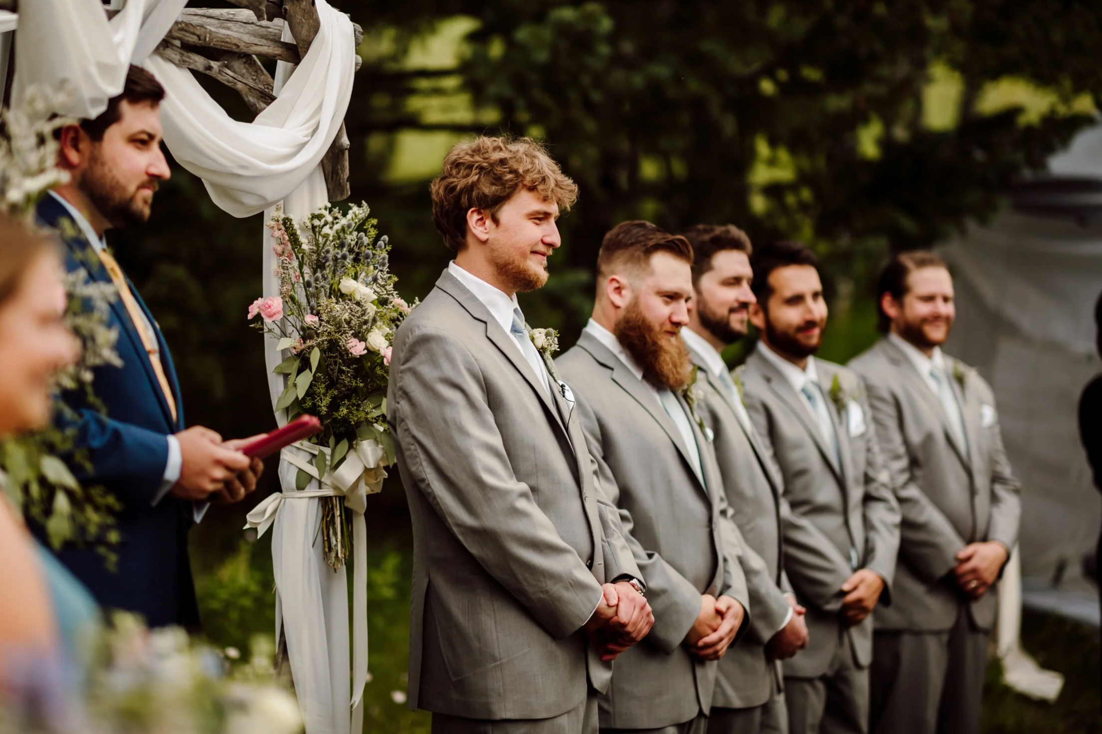A group of men dressed in suits standing outdoors at a wedding ceremony, with greenery in the background.