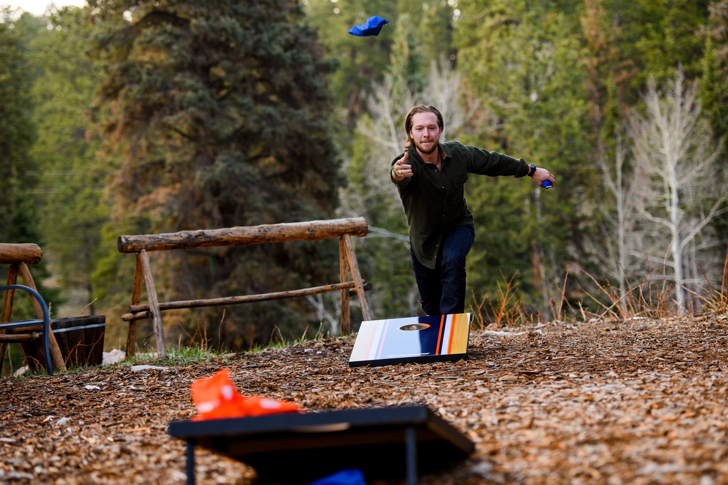 A man playing cornhole outdoors in a wooded area during daytime, with trees in the background.