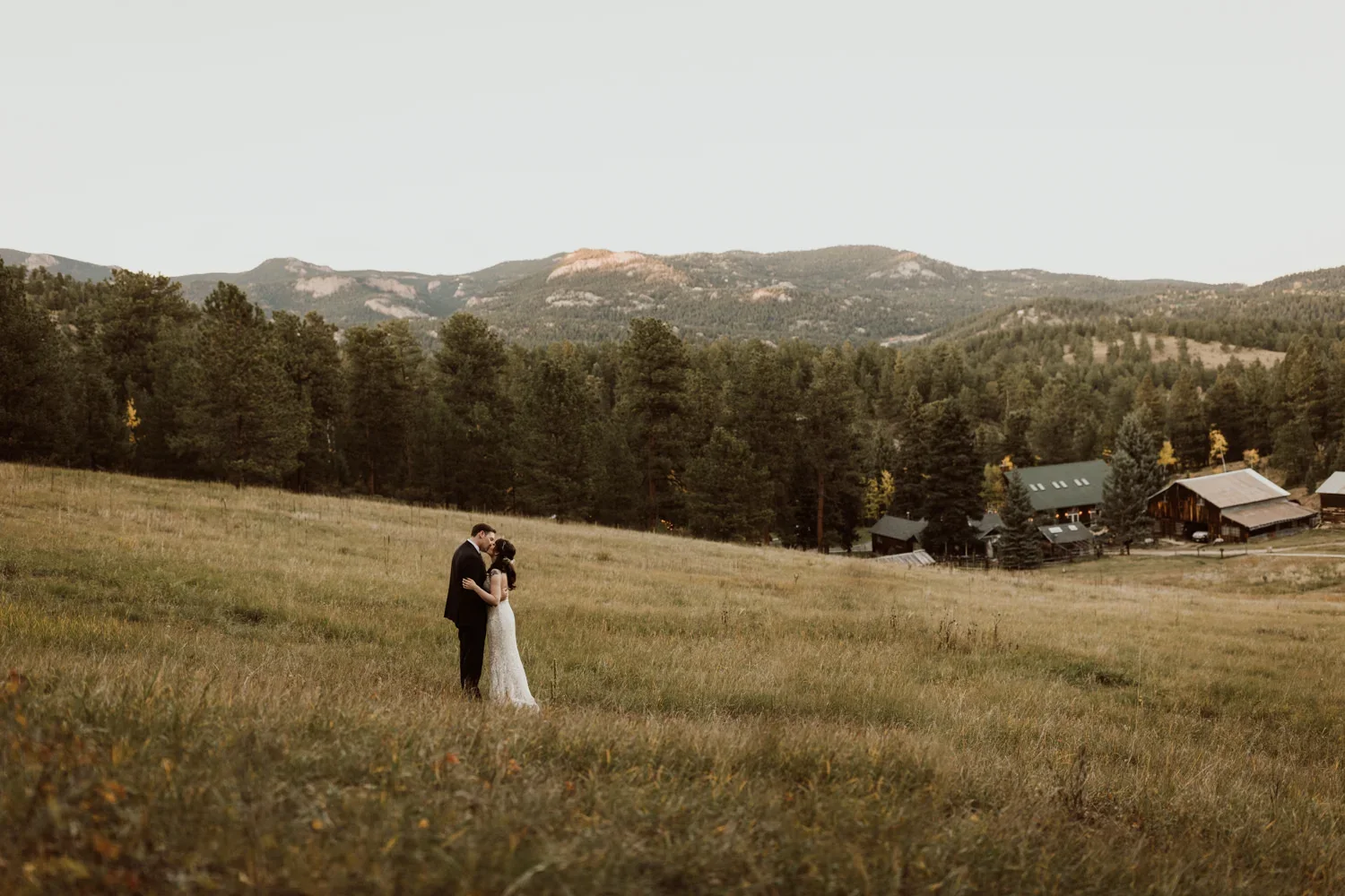 A bride and groom standing in a grassy field at sunset, embracing and kissing with mountains and trees in the background.