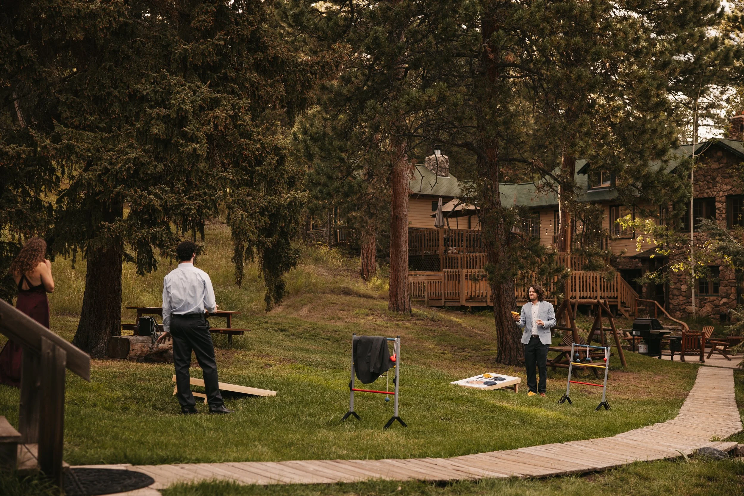People gathered outdoors in a backyard surrounded by tall trees, with a house visible in the background. Some individuals are standing, and there are outdoor games set up, such as cornhole. The scene appears to be a casual social gathering or outdoor party in the evening.