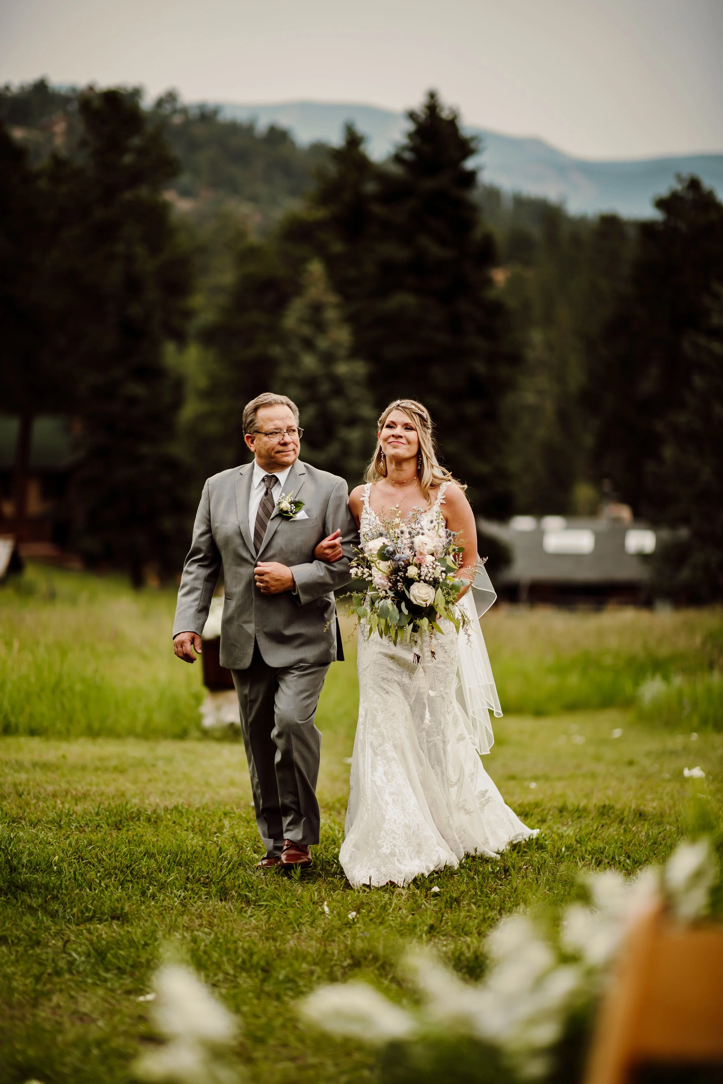 A bride in a white wedding dress with lace details and a bouquet walking down the aisle with her father at outdoor wedding ceremony with trees and mountains in the background.