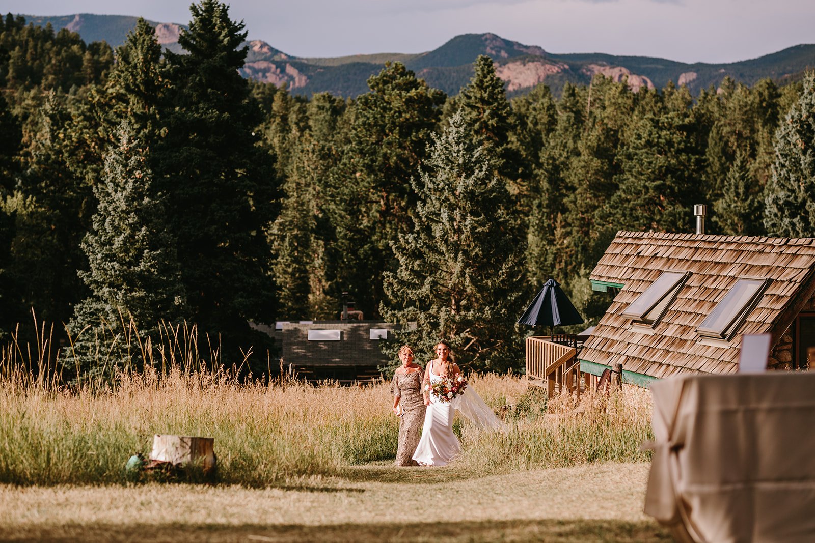 A bride and her mother walking in a grassy outdoor area, one in a wedding dress holding a bouquet, surrounded by trees and mountain scenery.