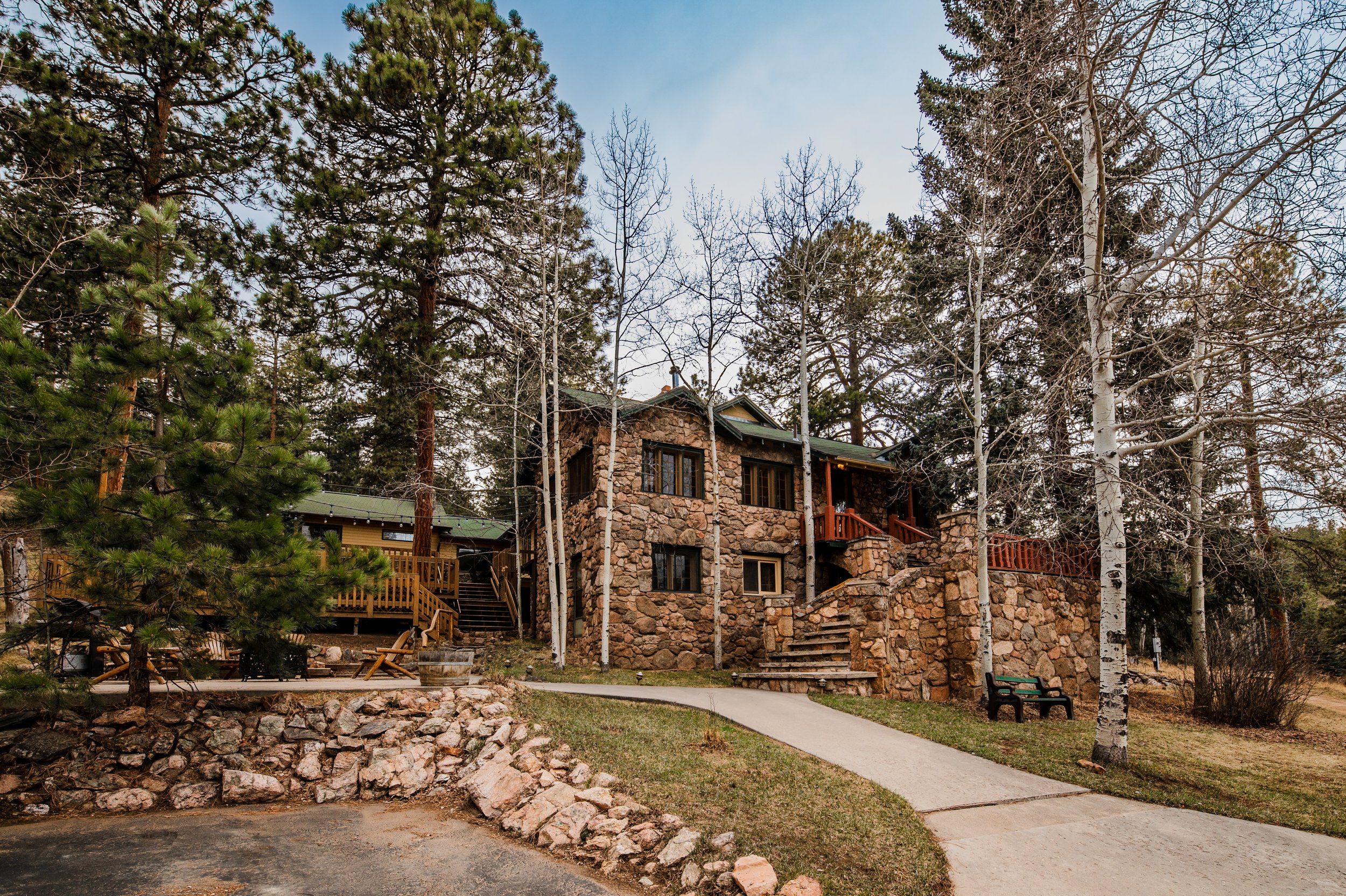 A stone house surrounded by tall trees, some with leaves and some without, with a paved walkway and benches in the yard.