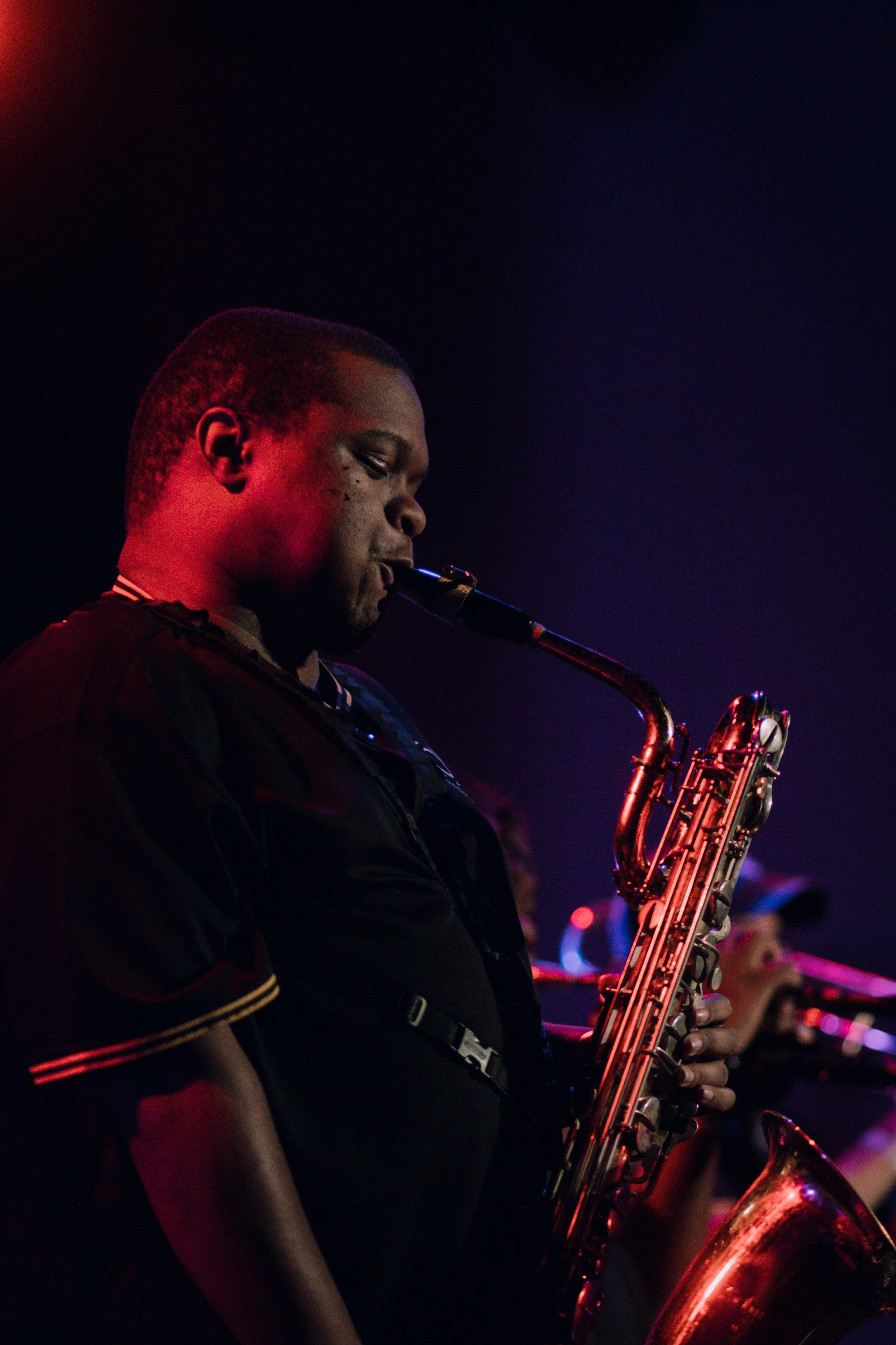 Dwayne Williams Jr of Emmanuel Ohemeng 3rd and Perpetual Motion Band playing a saxophone during a nighttime performance, with dark background and colorful stage lighting. (credit: @Soigné.Shutters)