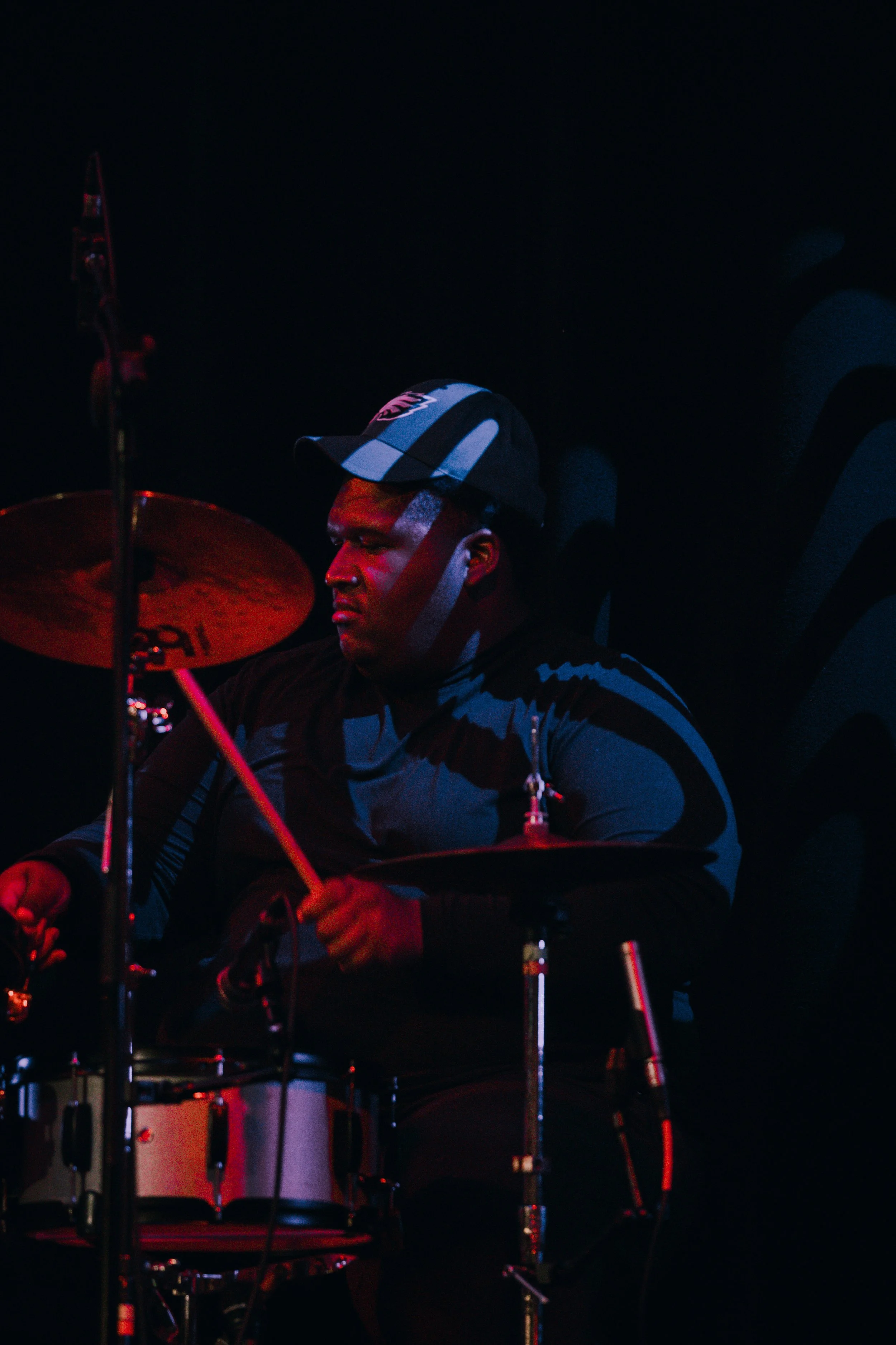 Jeremy "Caleb" Reynolds of Emmanuel Ohemeng 3rd and Perpetual Motion Band wearing a green Philadelphia Eagles cap playing a drum set in a dark room with colored lighting.