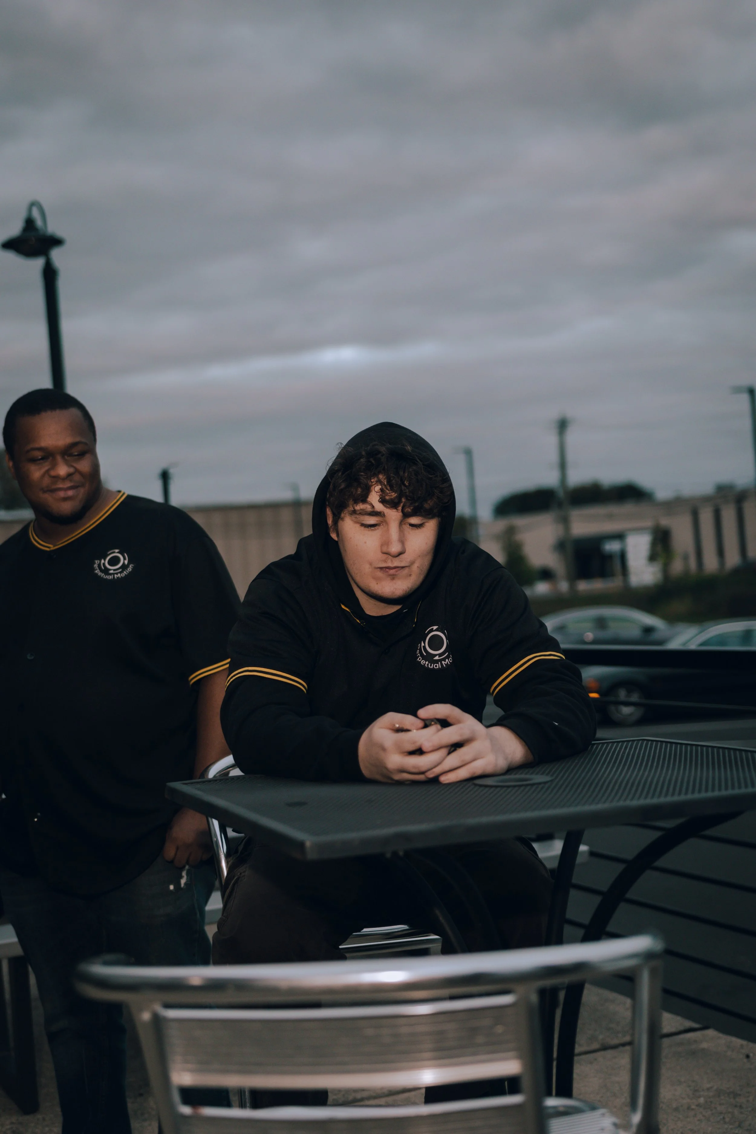 Two young men outdoors, one with curly hair wearing a hoodie, and the other with short hair, with overcast sky and park lights in the background.