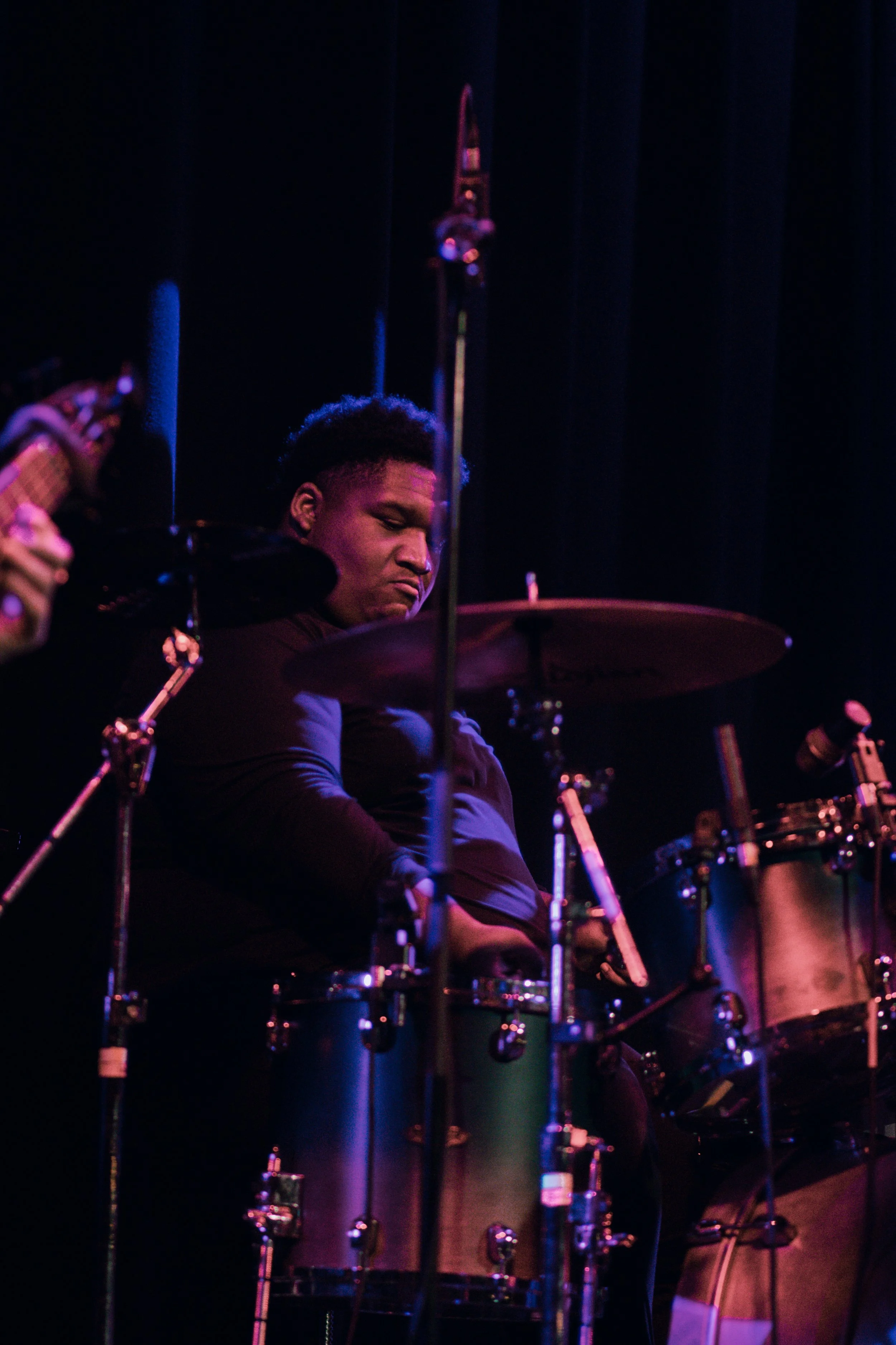 Jeremy "Caleb" Reynolds of Emmanuel Ohemeng 3rd and Perpetual Motion Band playing a drum set on stage with a dark background, illuminated by blue and purple stage lights.
