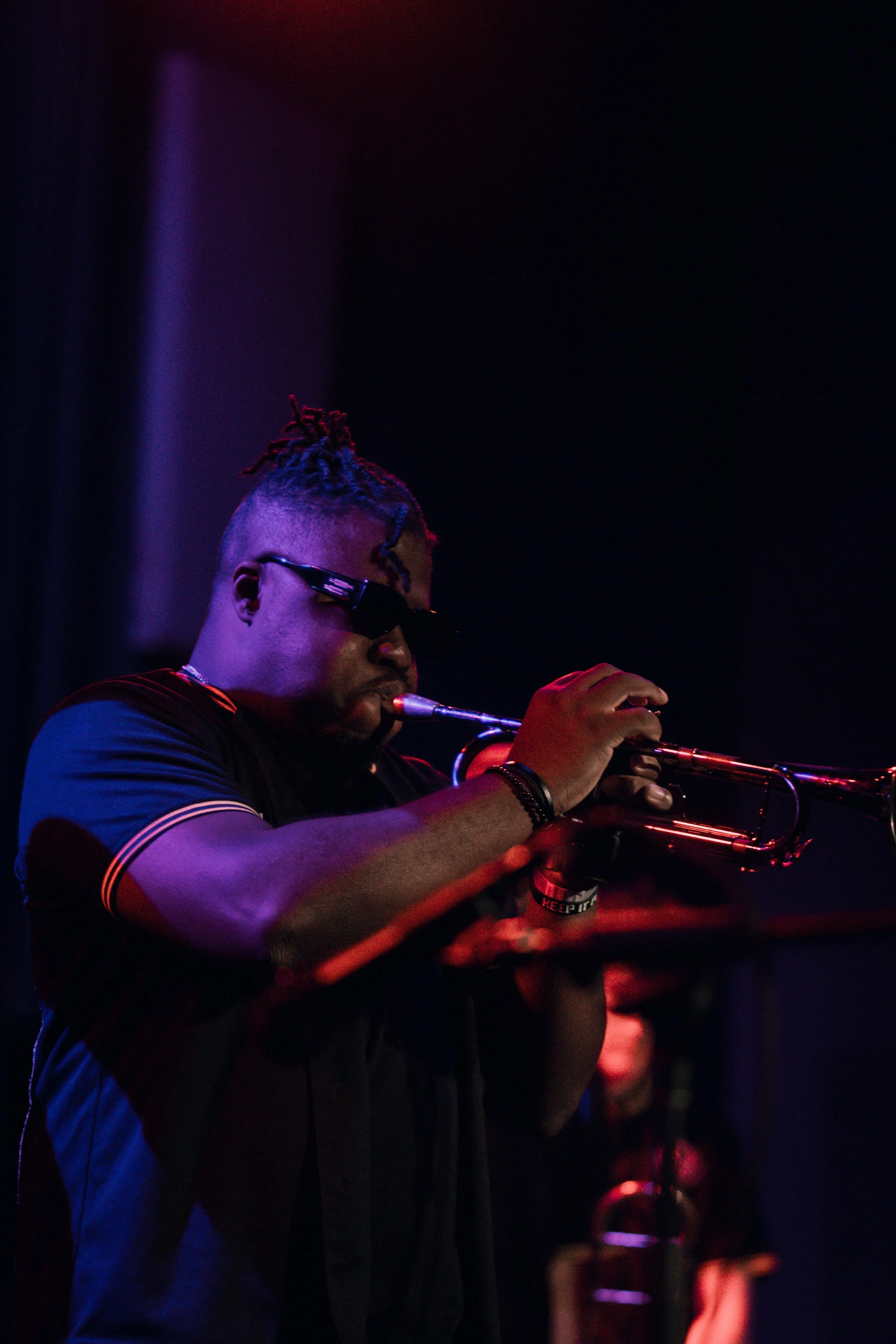 Emmanuel Ohemeng 3rd with dark sunglasses and dreadlocks playing a trumpet in a dimly lit setting with colorful lighting. (credit: @Soigné.Shutters)
