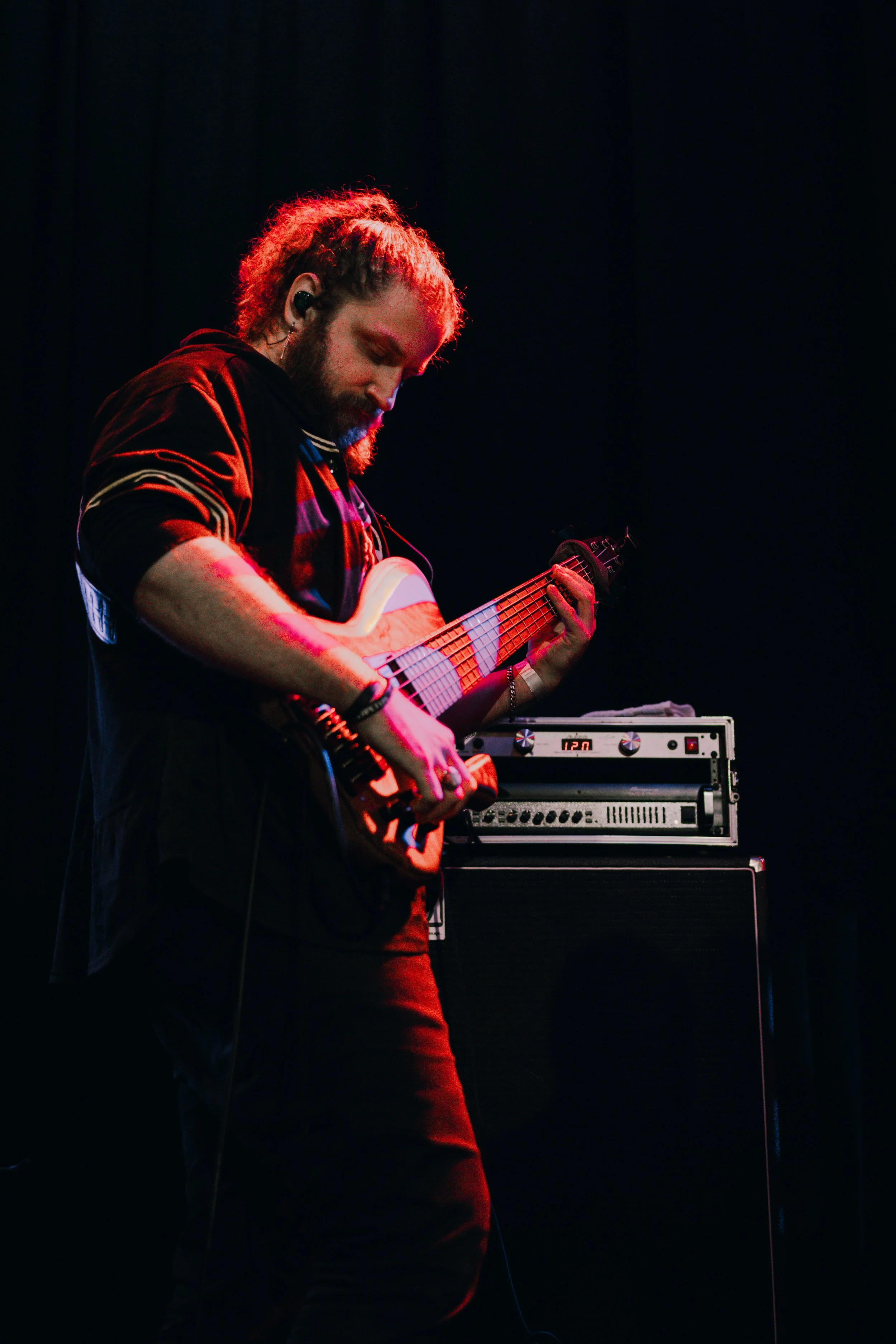 Christian Hencek of Emmanuel Ohemeng 3rd and Perpetual Motion Band with curly hair and a beard playing an electric guitar on stage, with professional audio equipment in the background, under dark lighting with red and blue hues.