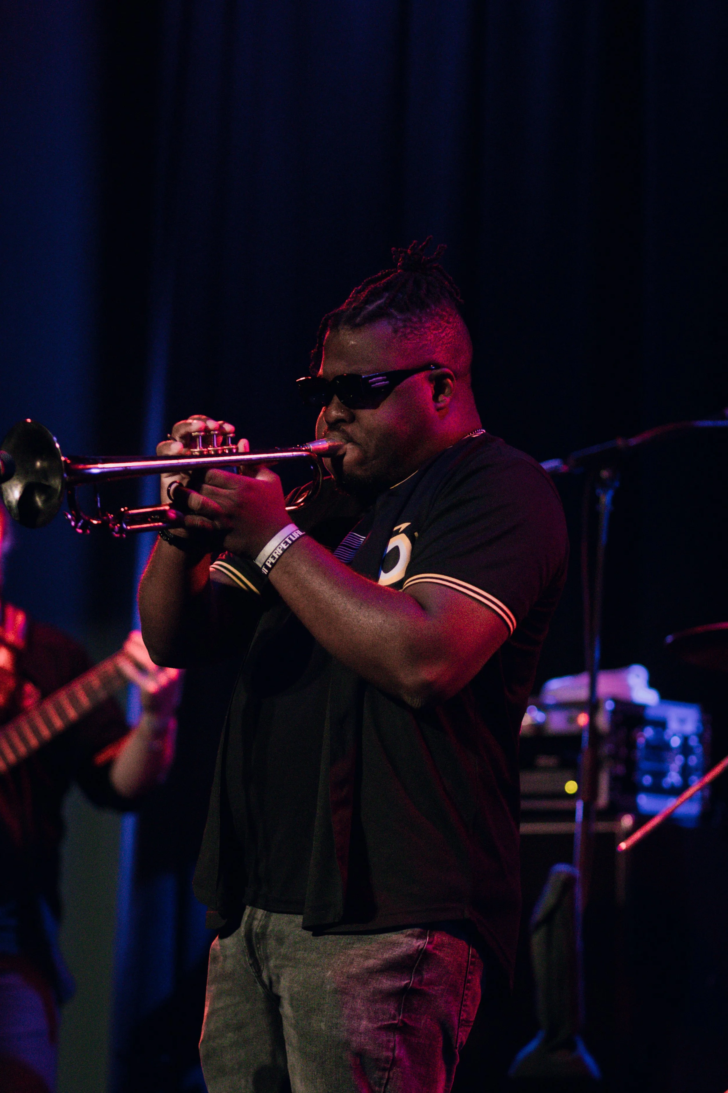 Emmanuel Ohemeng 3rd playing trumpet on stage, wearing sunglasses, a black shirt, and gray jeans, surrounded by dark blue stage lighting and other musicians.