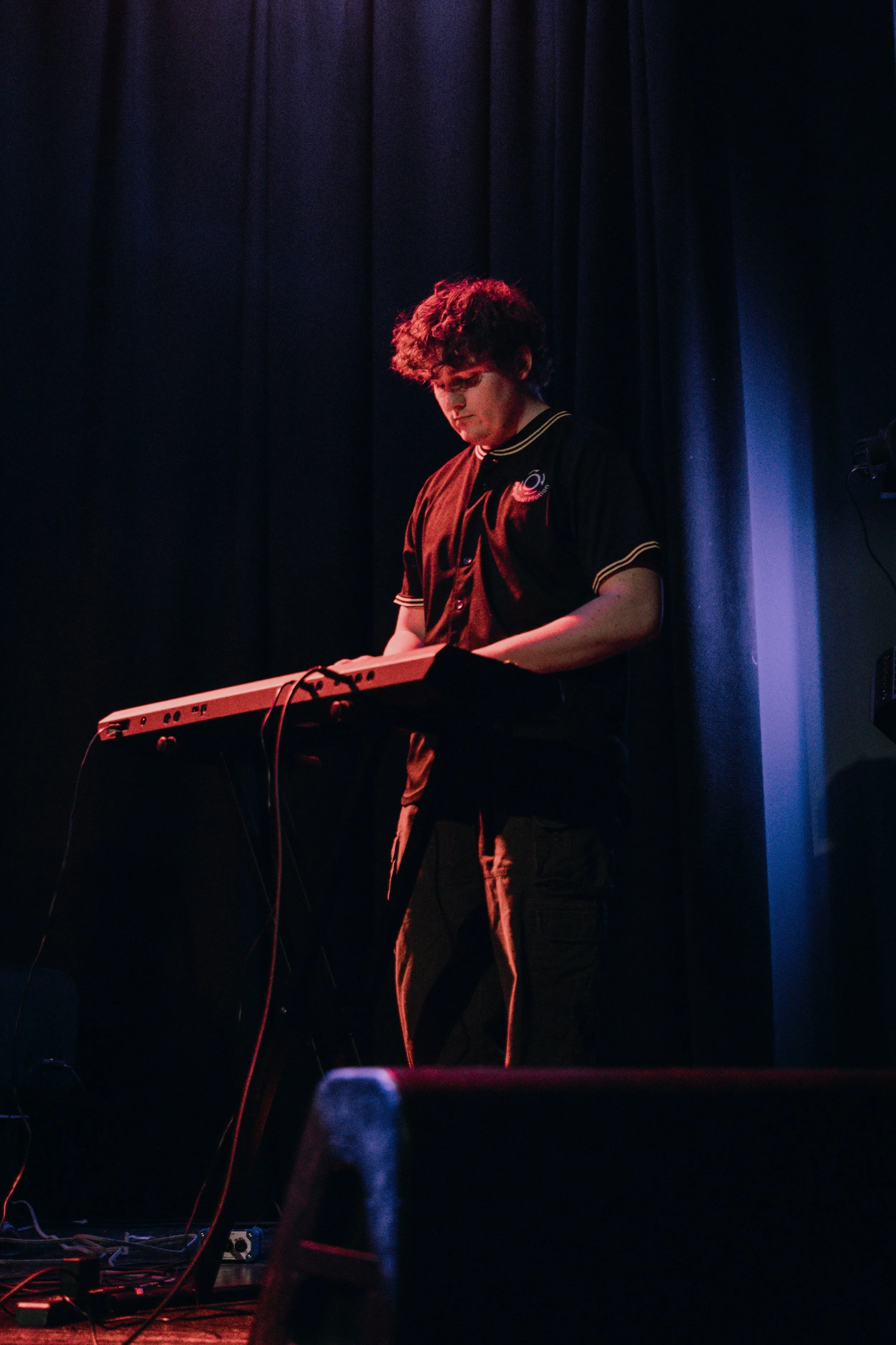 William McDonnell of Emmanuel Ohemeng 3rd and Perpetual Motion Band with curly hair playing a keyboard on stage during a live performance, dark background with stage lighting.