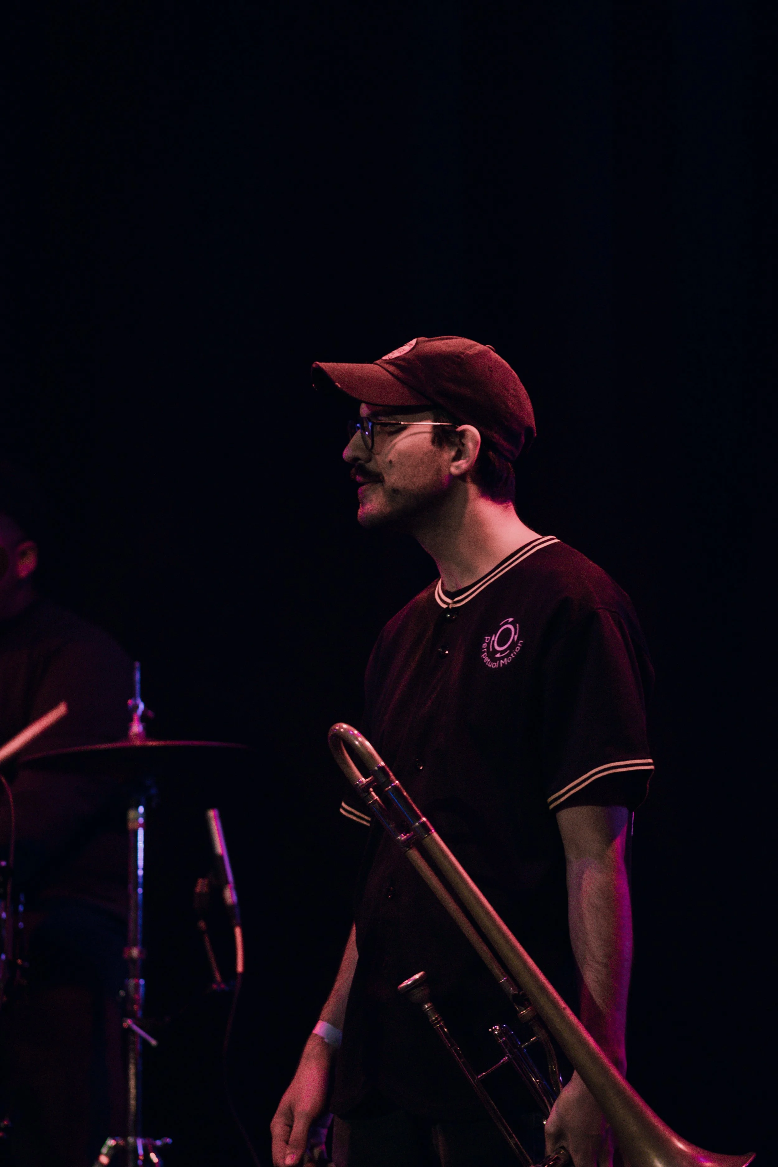 Bill Saurman of Emmanuel Ohemeng 3rd and Perpetual Motion Band wearing a red cap and glasses holding a trombone in a dark environment, illuminated by pinkish-purple lighting, possibly during a performance.  (credit: @Soigné.Shutters)