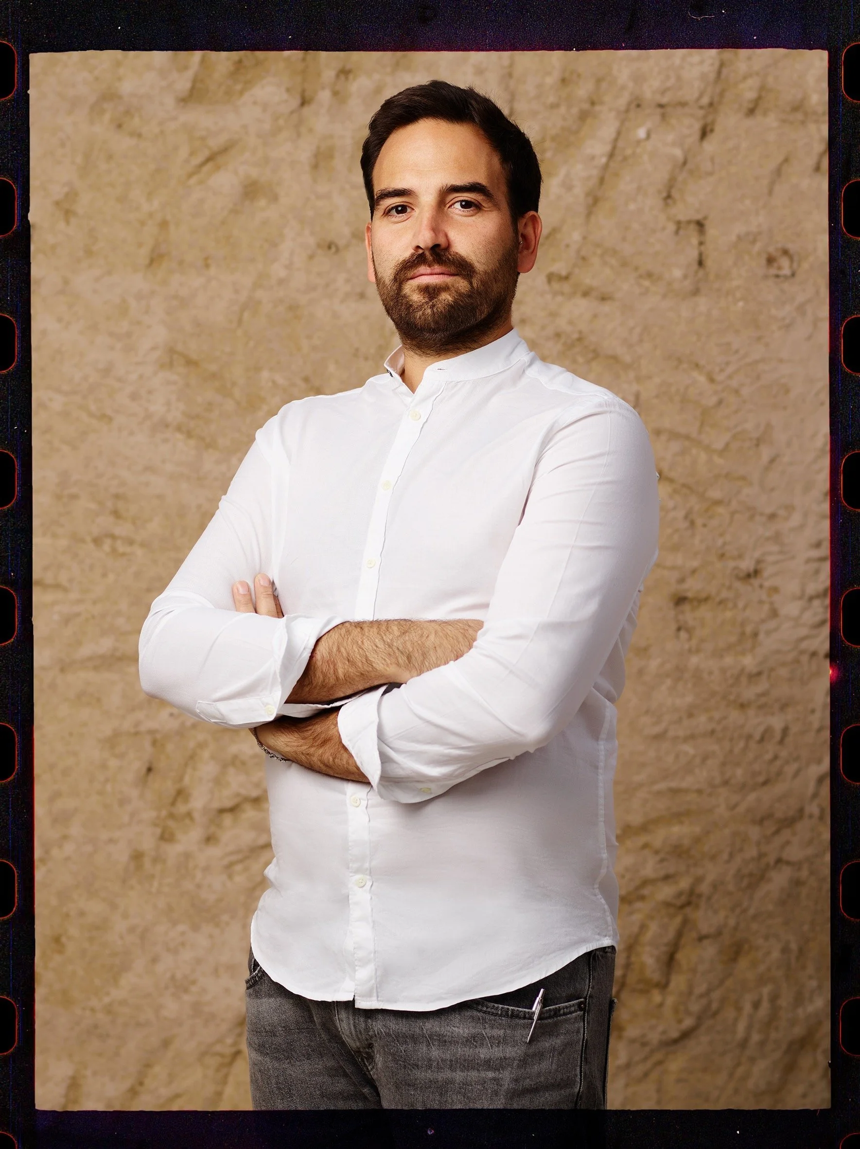 Uomo con barba che indossa una camicia bianca con le braccia incrociate, sfondo beige.