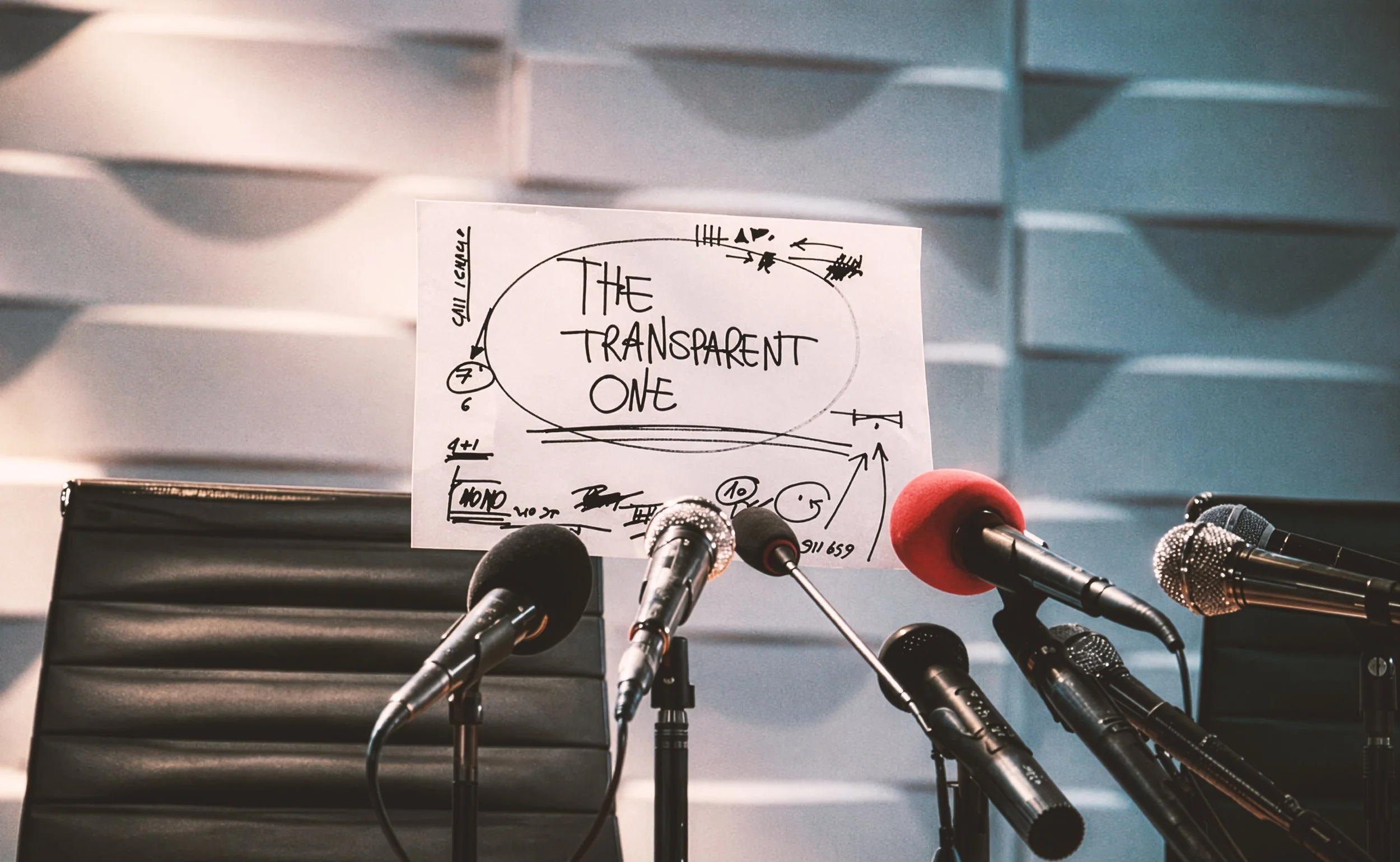 Una conferenza stampa con microfoni disposti su un tavolo in primo piano e un cartellone con scritto 'The Transparent One' sullo sfondo.