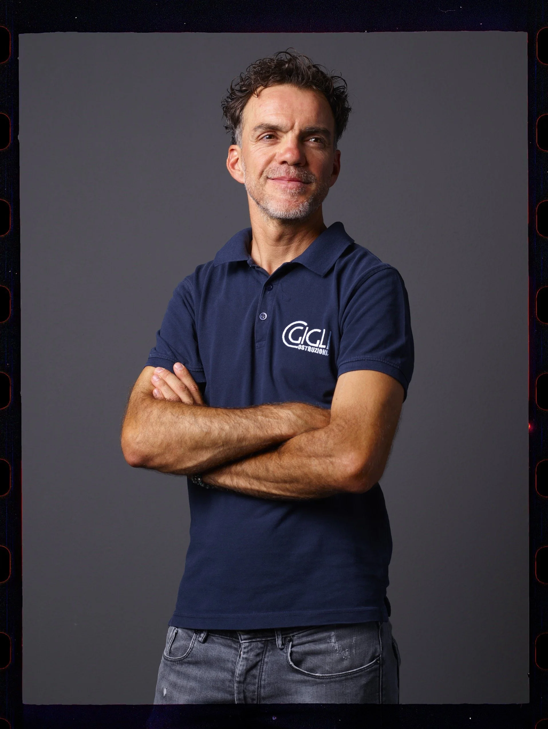 Ritratto di un uomo con capelli ricci e barba, indossa una polo blu con logo GCL e jeans grigi, sfondo grigio