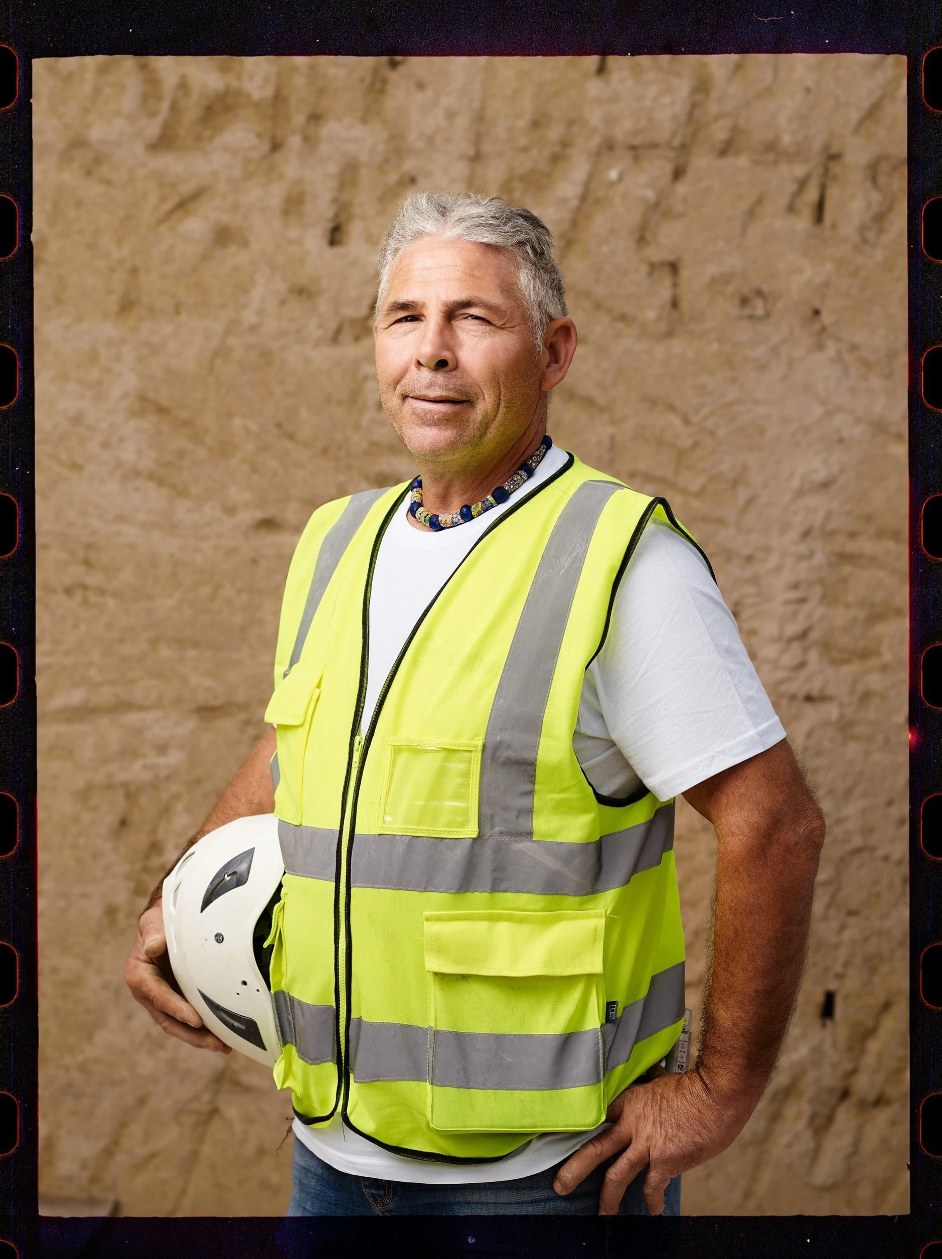 Uomo con capelli grigi, senza maglietta, che indossa un giubbotto di sicurezza giallo con strisce riflettenti, tiene in mano un casco bianco con visiera e porta una collana di perline colorate. Sfondo di muro in pietra.