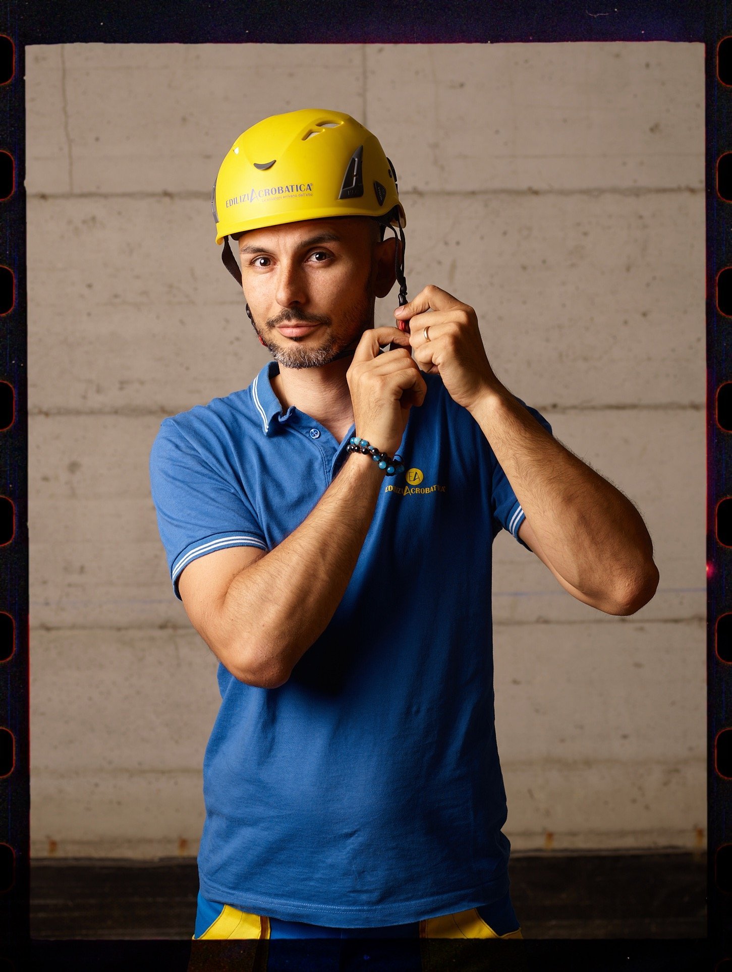 Uomo che indossa un casco giallo e una maglietta blu, che si sta preparando per un'attività di arrampicata o avventura, in un ambiente interno con parete di cemento.