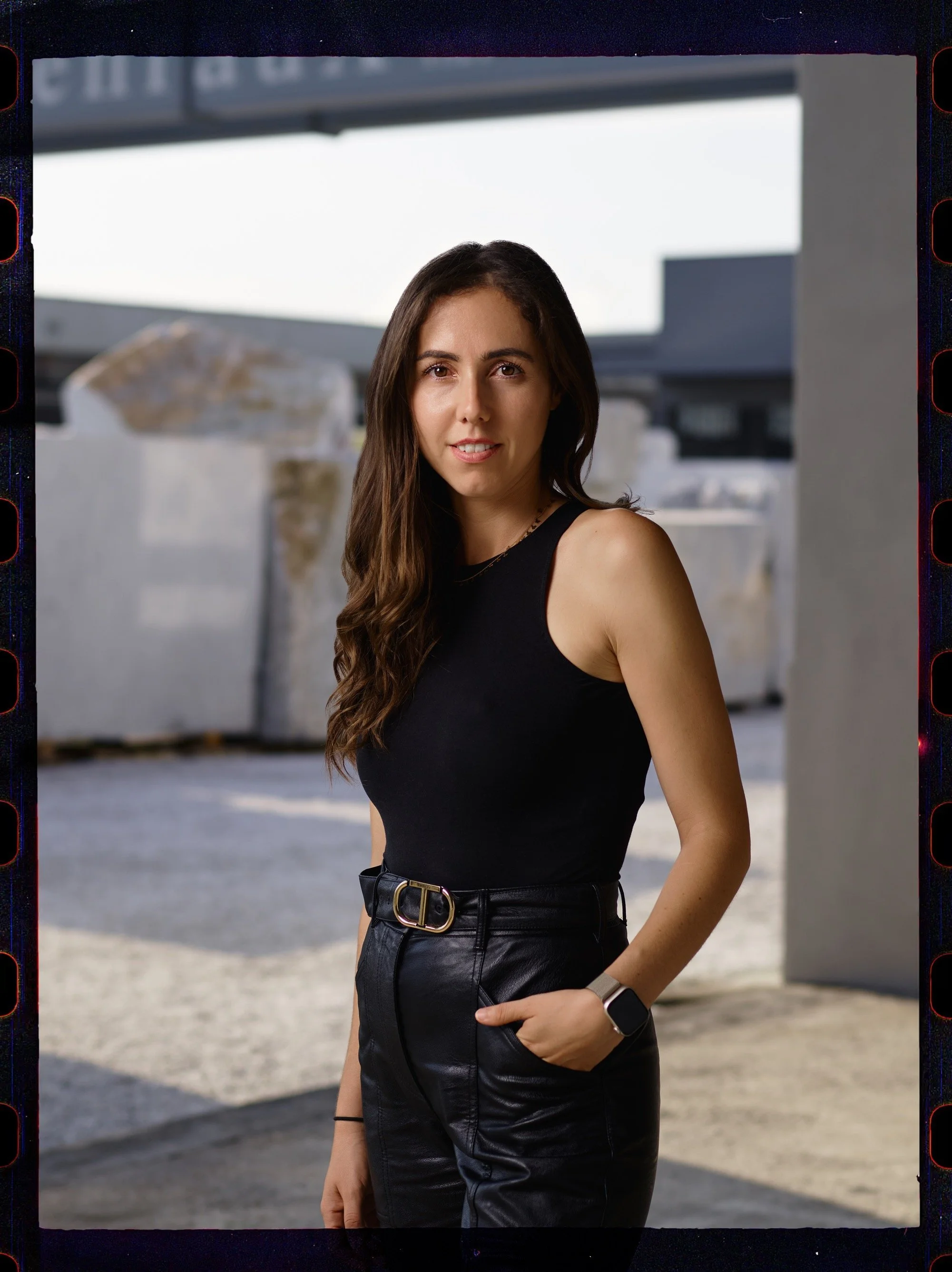 Una giovane donna con capelli lunghi e ricci, indossa una maglietta nera e pantaloni in pelle neri con cintura argento, e ha un orologio smartwatch sul polso, si trova all'aperto con uno sfondo di strutture architettoniche moderne.