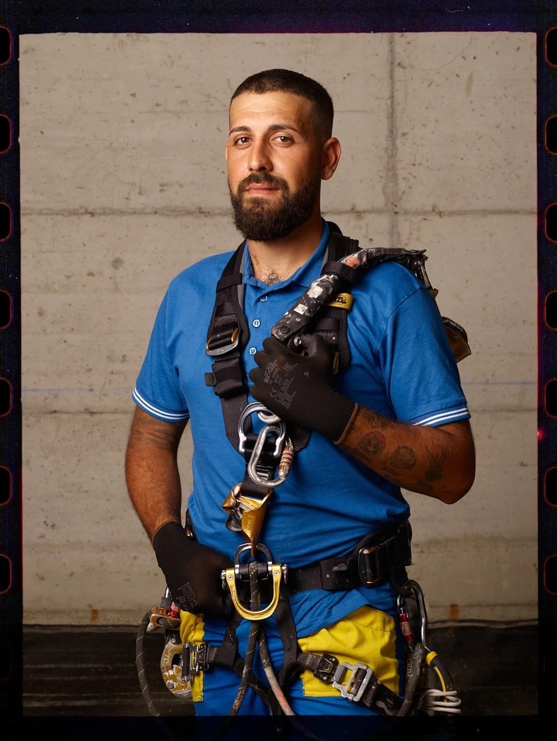 Uomo vestito con abbigliamento da lavori in quota, con imbracatura di sicurezza e ferramenta, posa davanti a un muro in cemento.