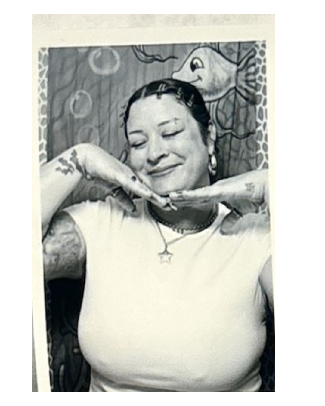 A black-and-white photo of a woman with tattoos, smiling with her eyes closed, posing with her hands under her chin; background features a colorful artwork of a fish.