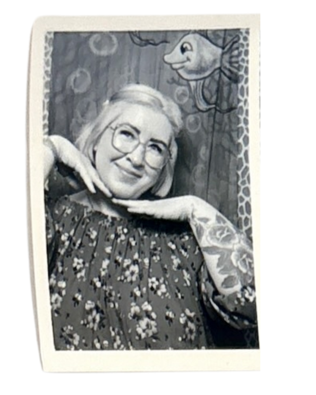 Black and white photo of a woman with glasses smiling, posing with her hands under her chin. She has floral tattoos on her arm and is dressed in a floral patterned top. A cartoonish fish with a happy face is in the background, painted or illustrated on a wooden surface.
