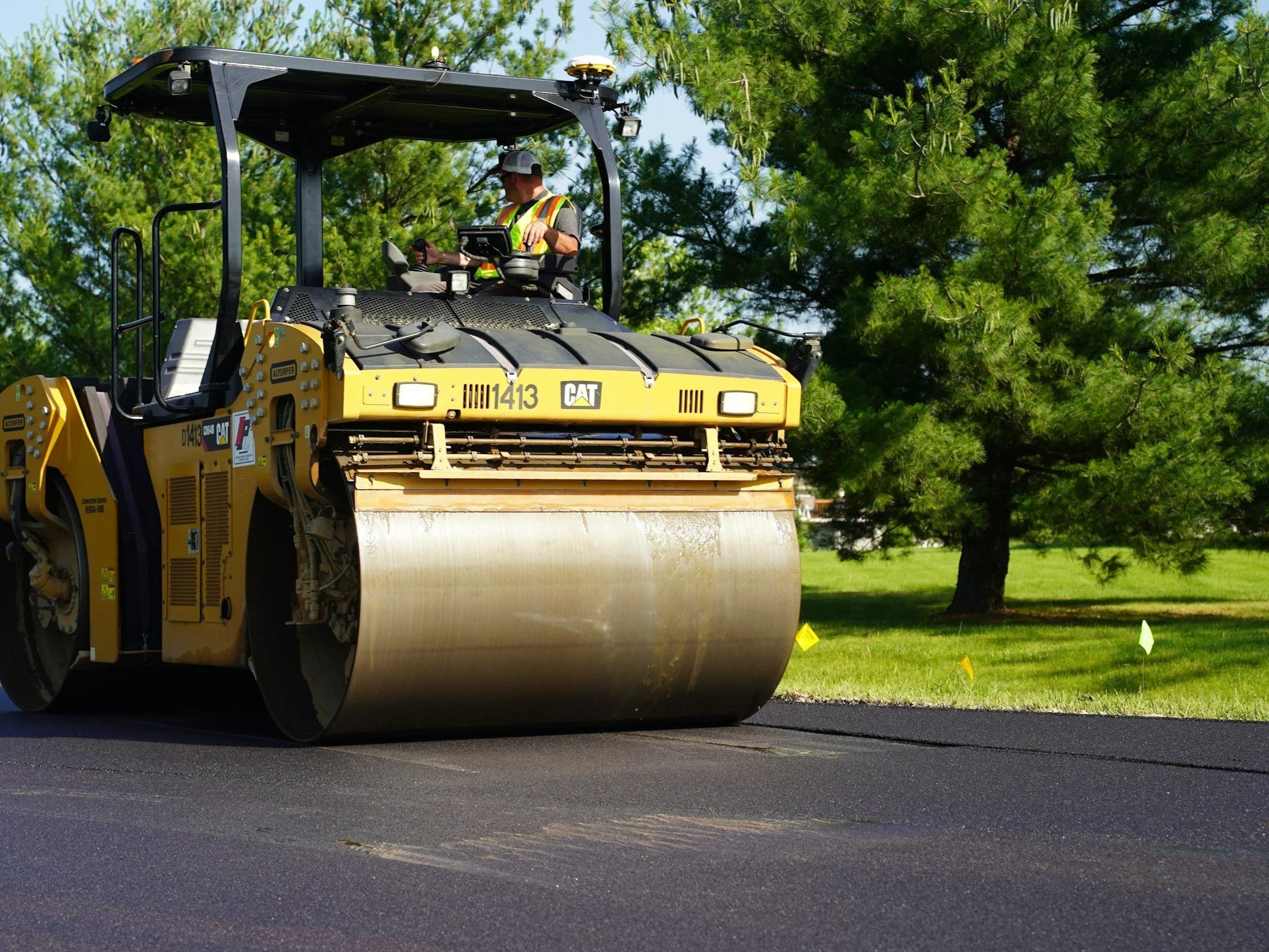 A worker operating a yellow CAT construction roller machine on a freshly paved surface, surrounded by green trees and a partly cloudy sky in the background.