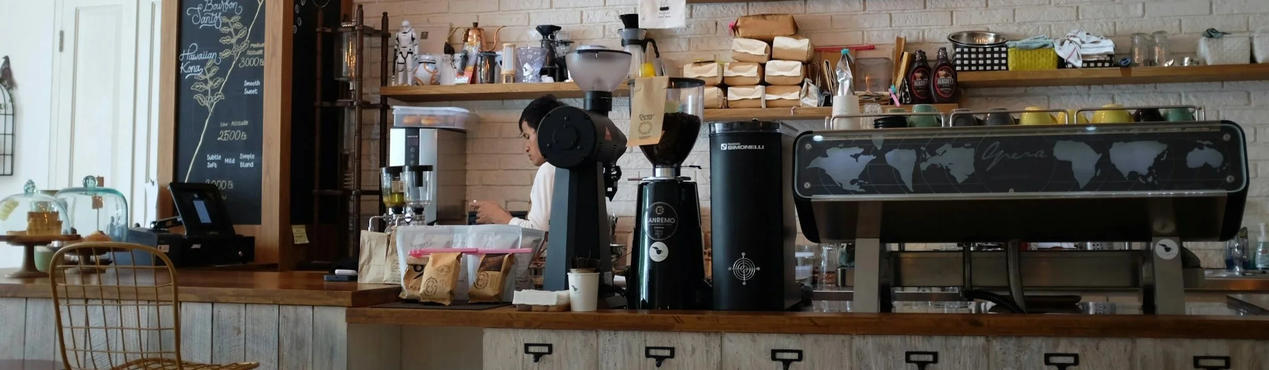 A barista preparing drinks behind the counter in a cafe, with coffee-making equipment, a laptop, and paper bags visible on shelves, and a chalkboard menu listing different flavors and prices.