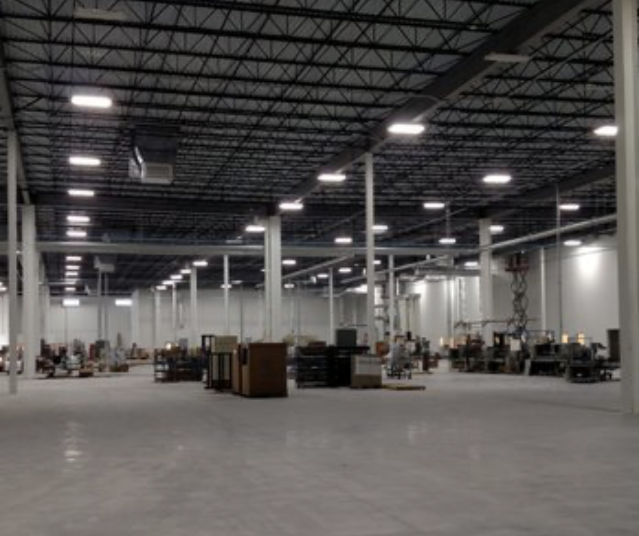 Empty warehouse with high ceilings, industrial lighting, and scattered furniture and equipment.