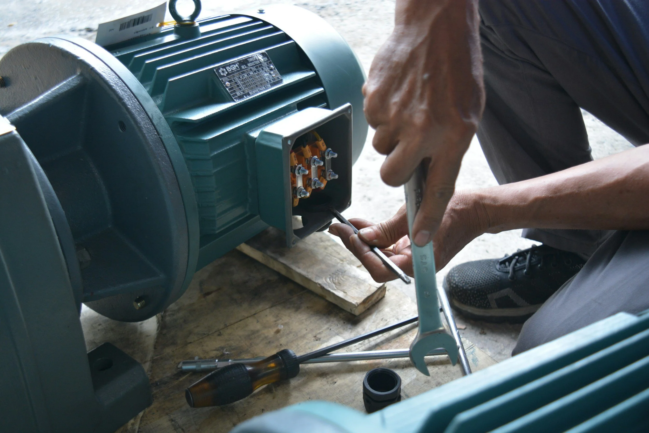 Person working with electrical motor, holding a wrench near the motor's terminal box, on a wooden surface, surrounded by tools.