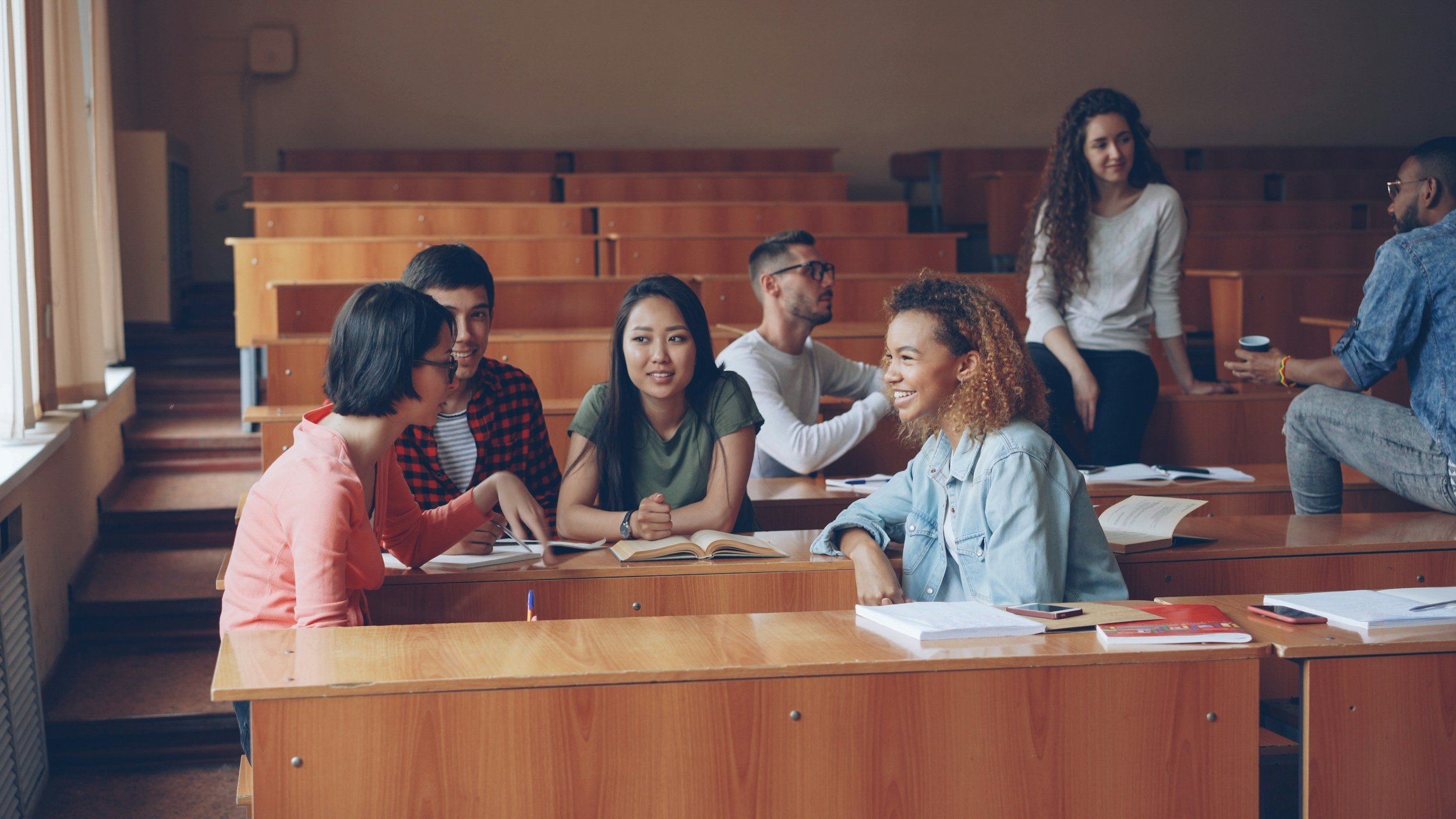 A group of college students sitting at wooden desks in a classroom, engaging in conversations and studying, with some standing and others seated.