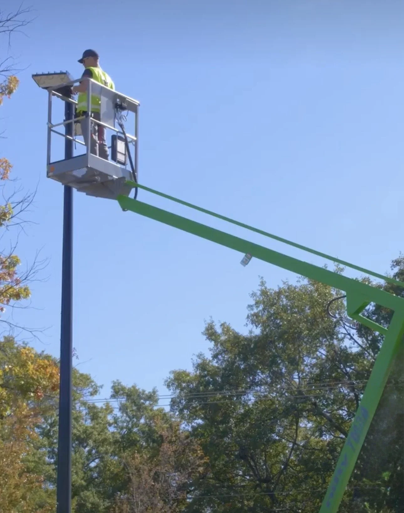 A worker in a yellow safety vest and dark hat operating a hydraulic lift, working on streetlights in a park with trees and a clear blue sky.