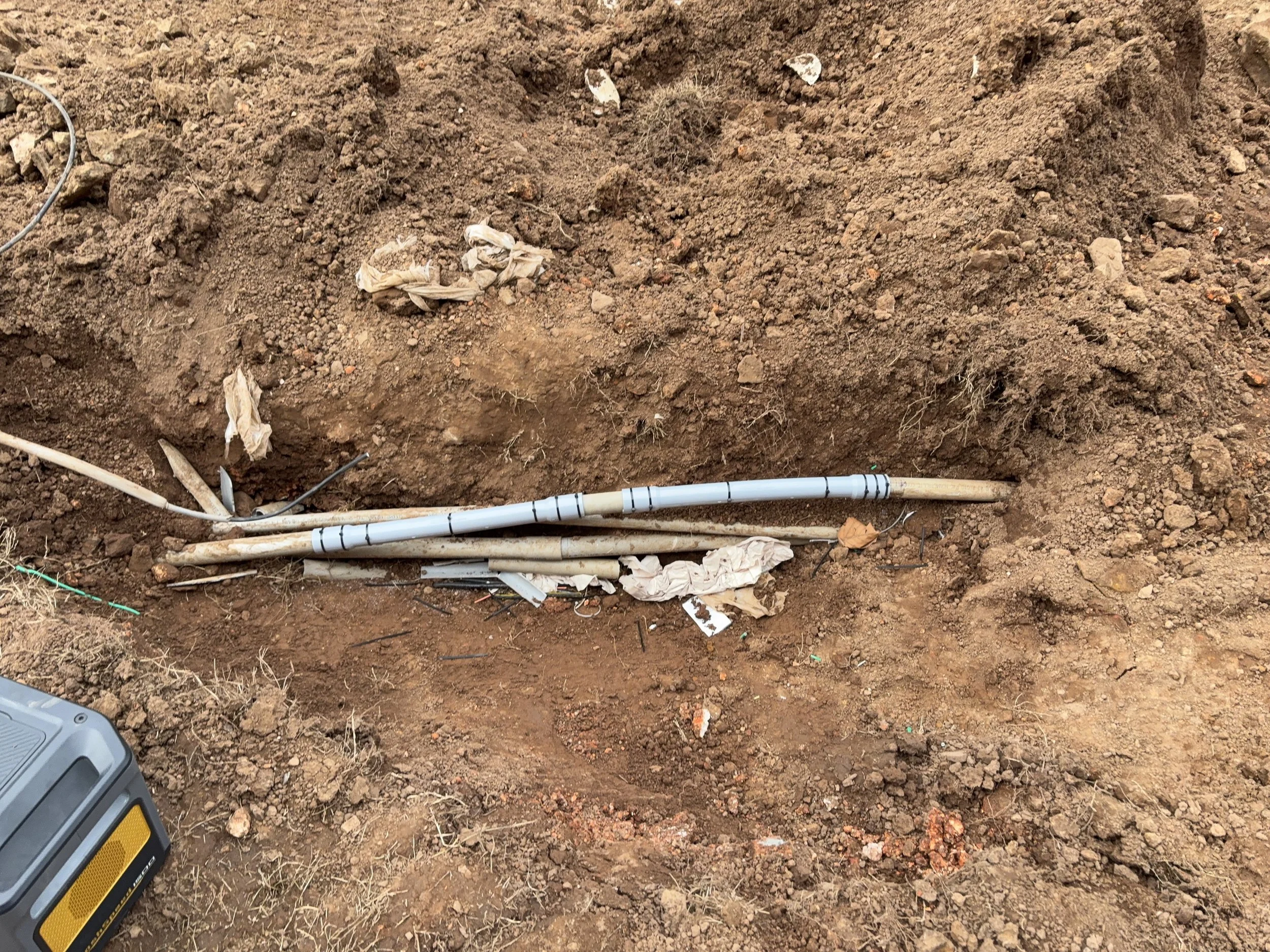 A trench in the ground with exposed dirt, pipes, and discarded paper debris.