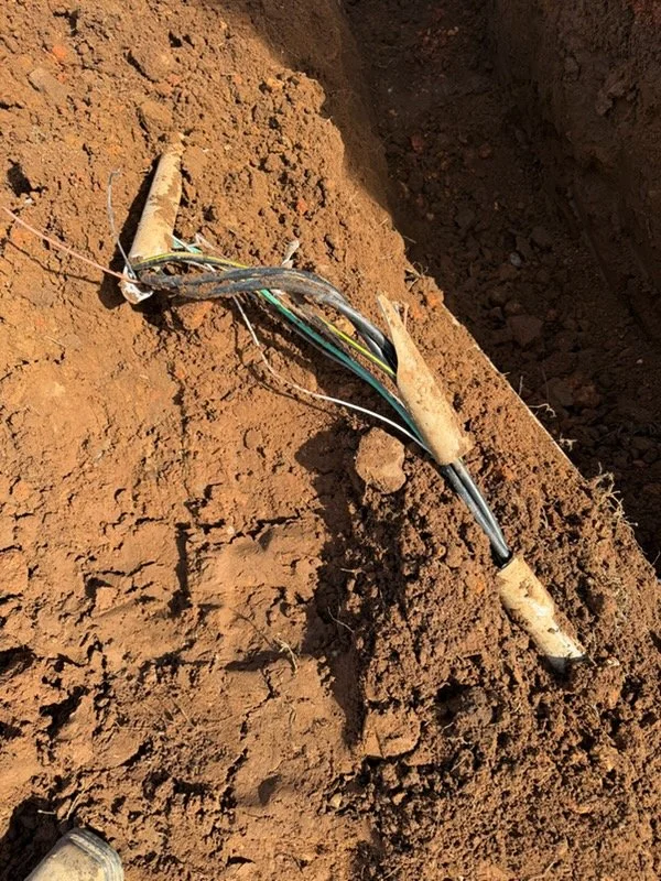Exposed electrical wires on red dirt, with some covered in white protective material, and a black cable connected to a black plastic junction box.