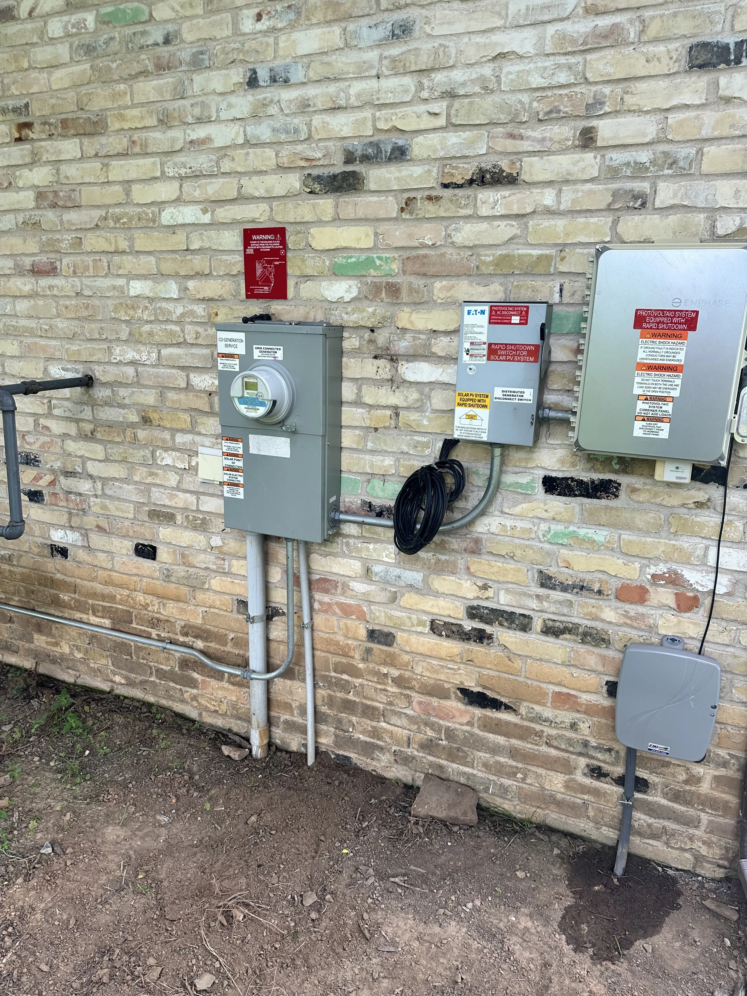 A brick exterior wall with various electrical and solar power system equipment fixtures, including circuit boxes, warning labels, and cables, mounted on the wall and connected to underground wiring.