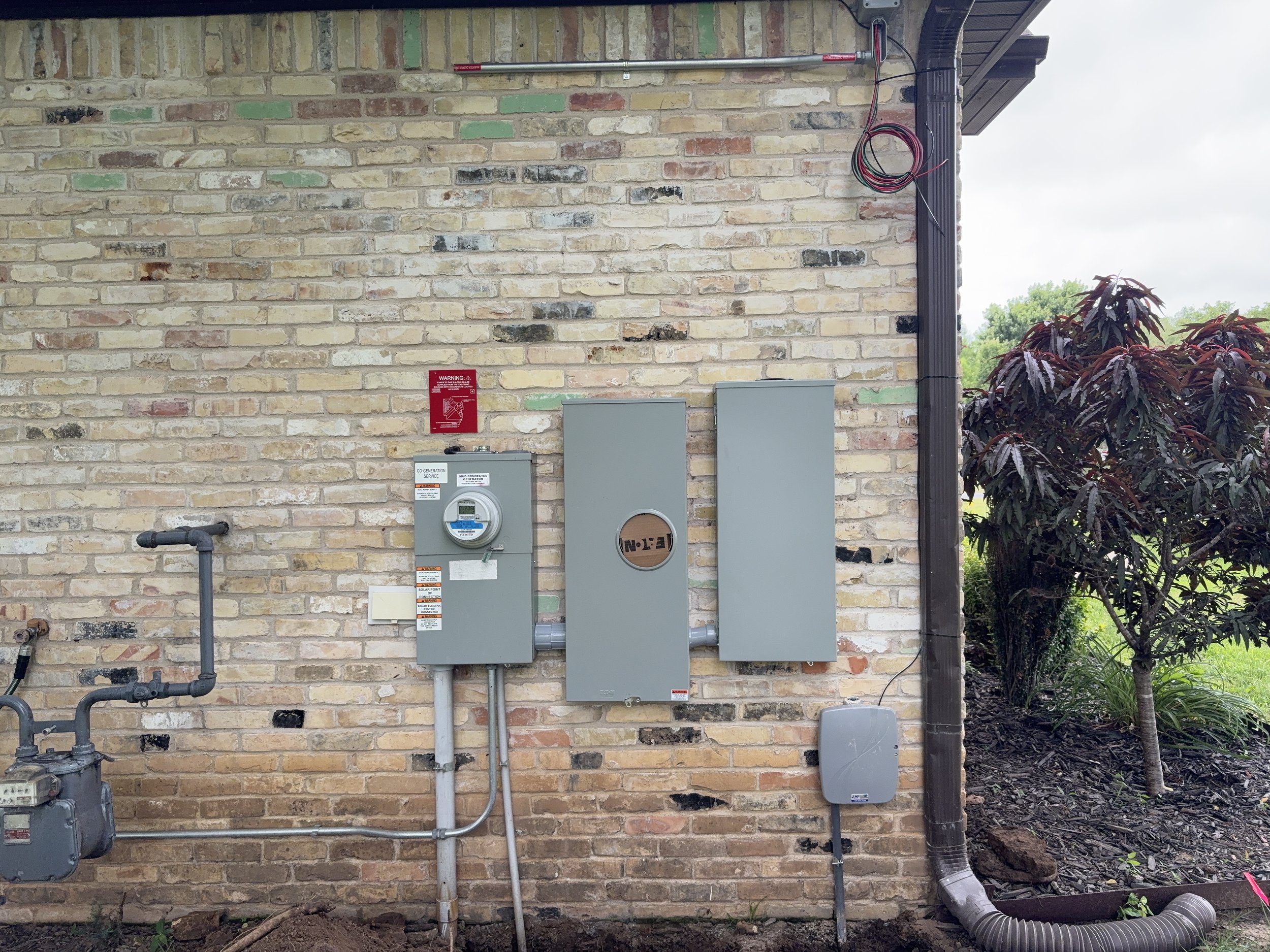 Exterior brick wall with electrical boxes, wiring, pipes, and a warning sign, with some trees and plants to the right.