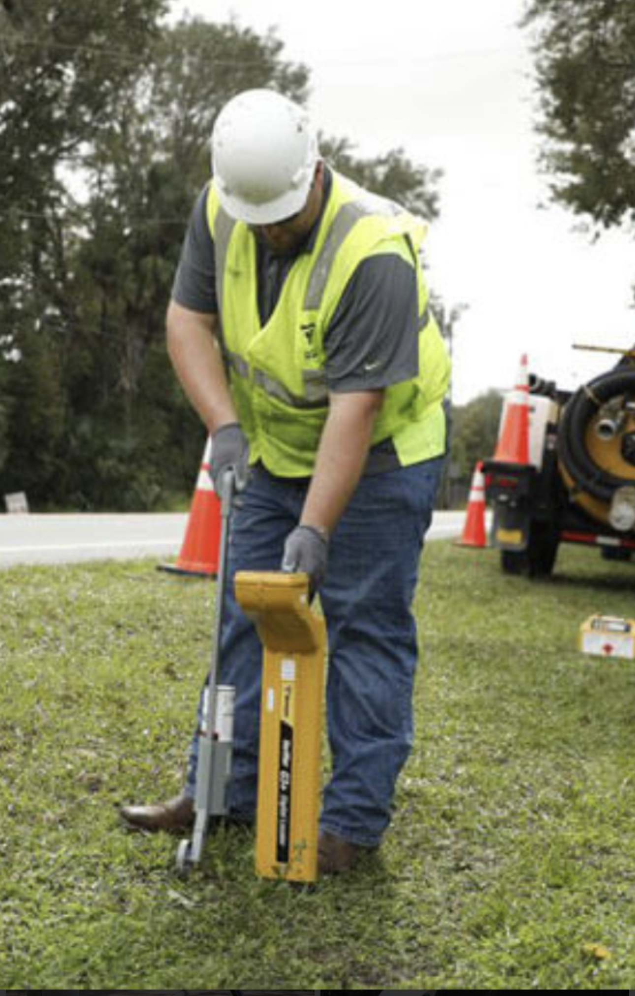Construction worker wearing a white hard hat, yellow safety vest, and gray shirt working with construction equipment on a grassy area, with orange traffic cones and trees in the background.