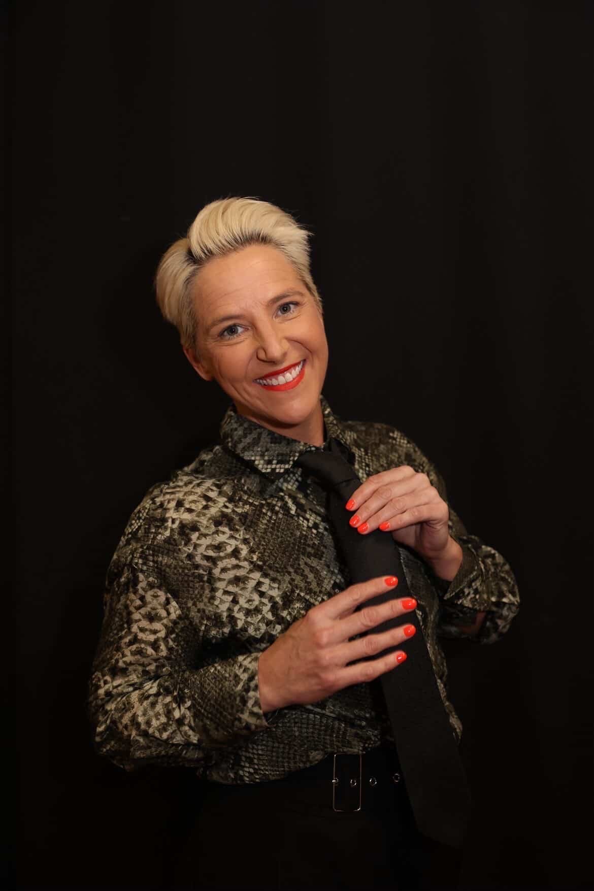 A woman with short platinum blonde hair, smiling, wearing a snake-print blouse, black tie, and bright red nail polish, standing against a black background.