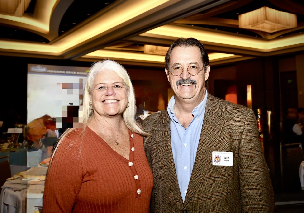 A smiling woman with long blonde hair and a man with glasses, a mustache, and brown hair, standing together in a conference room. The woman is wearing an orange top with buttons, and the man is wearing a brown blazer and a blue shirt with a name tag.